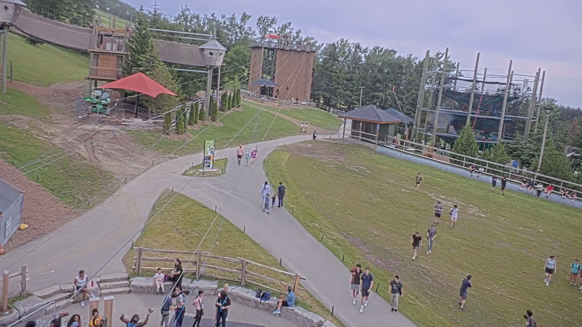 An outdoor recreational area with various structures, including climbing towers and zip lines, features several people walking on paved paths and grassy areas under an overcast sky.