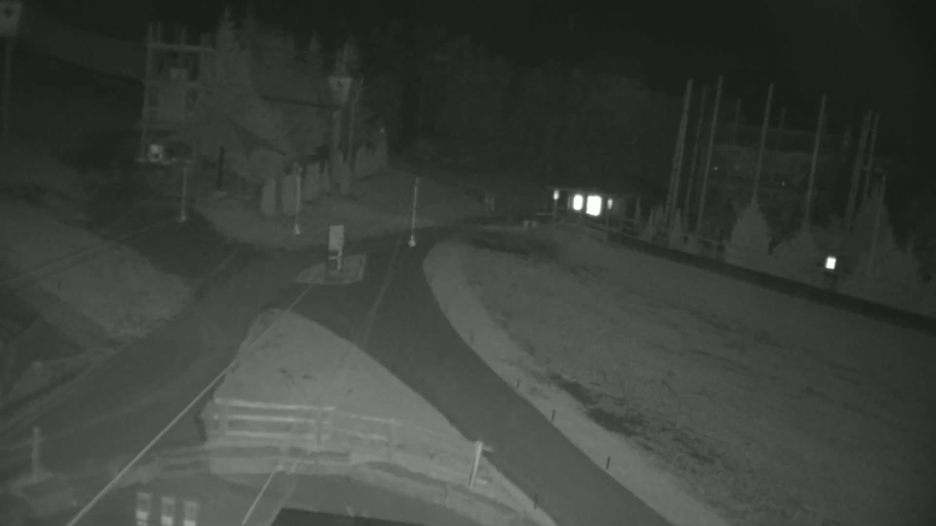 A nighttime, low-light view shows a paved road curving through a sparsely-lit area with structures and trees visible in the background.
