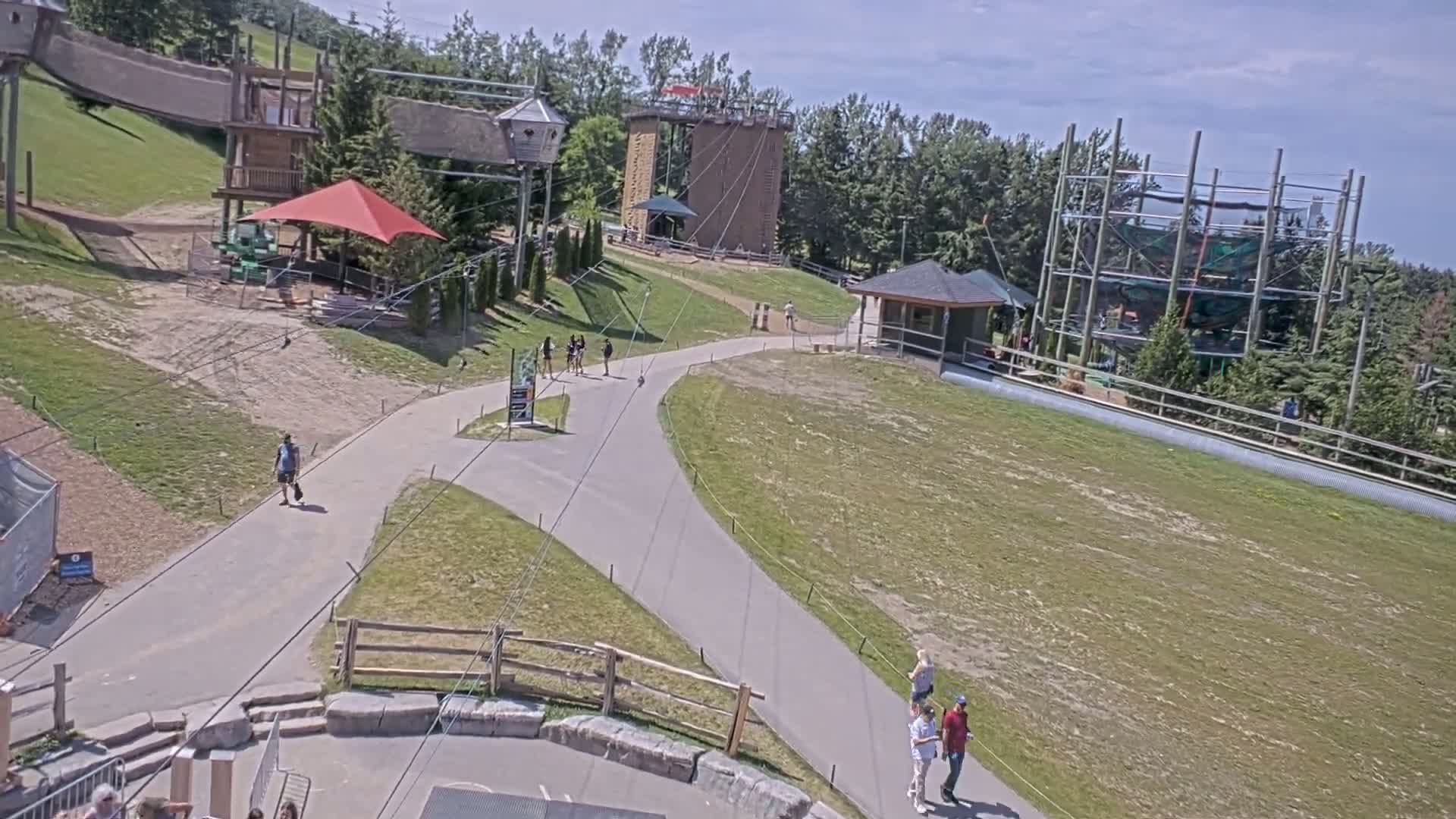 An outdoor recreational area with paved paths, various play structures, and a climbing wall under sunny skies.