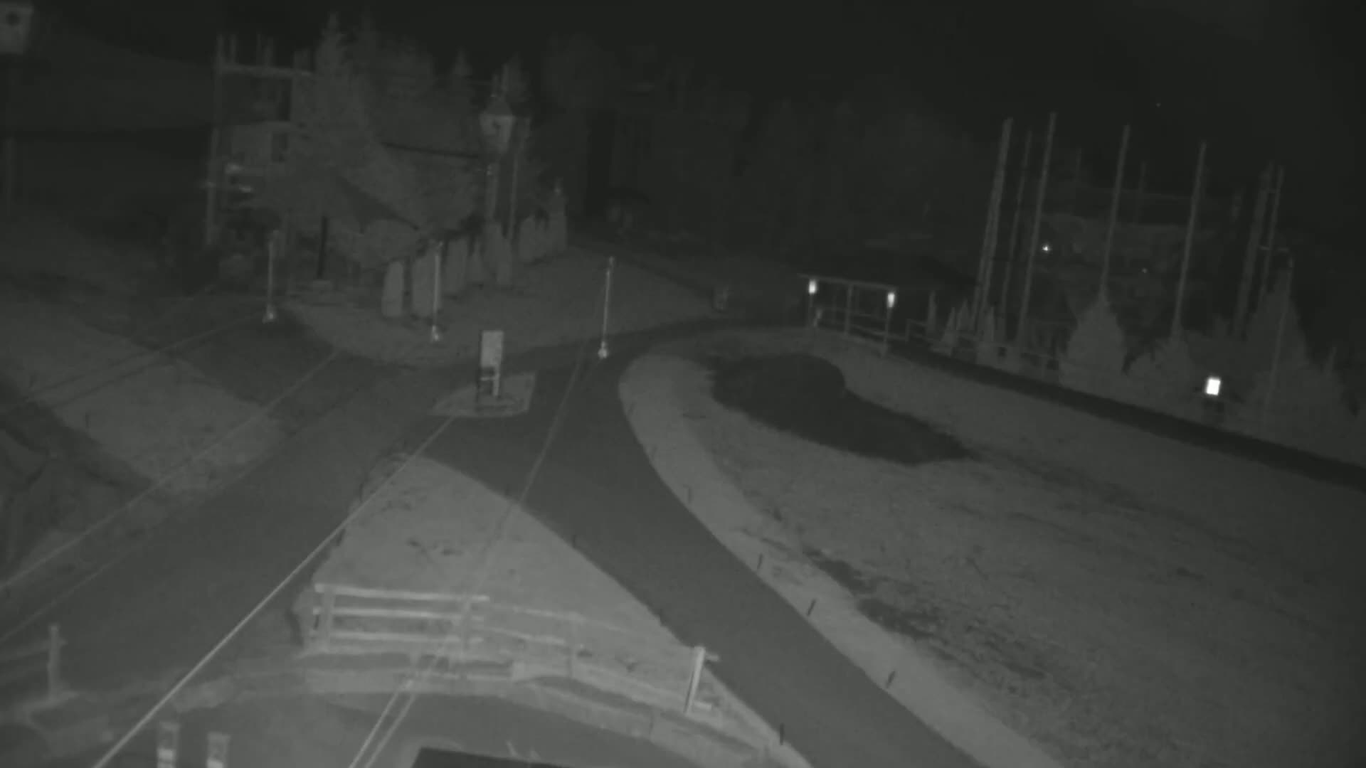 A nighttime, low-light view shows a paved area with a curved roadway and structures in the background.