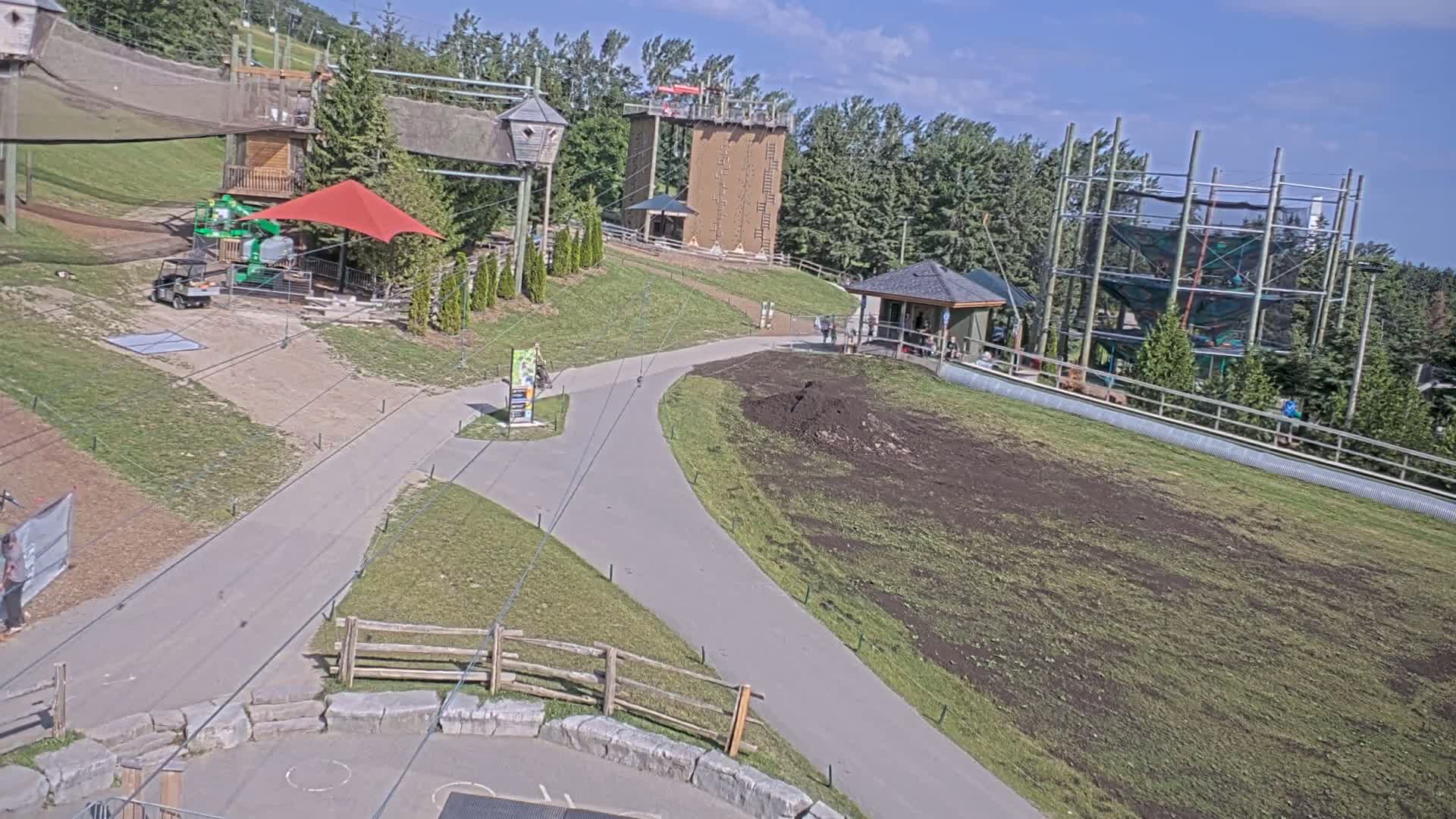 A paved pathway winds through a grassy area with various outdoor recreational structures, including climbing walls and a zip line course, under sunny skies.