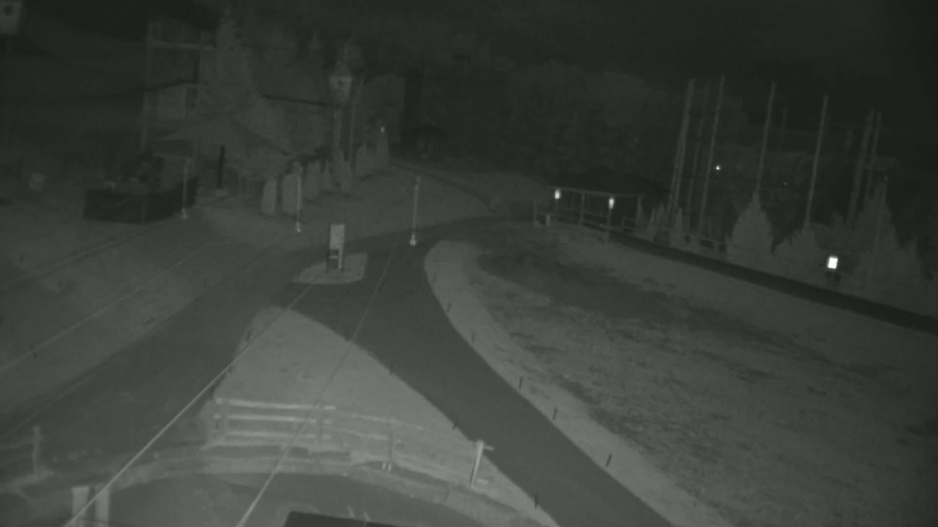 A nighttime, low-light image shows a paved road curving around a sparsely-lit area with indistinct structures in the background.