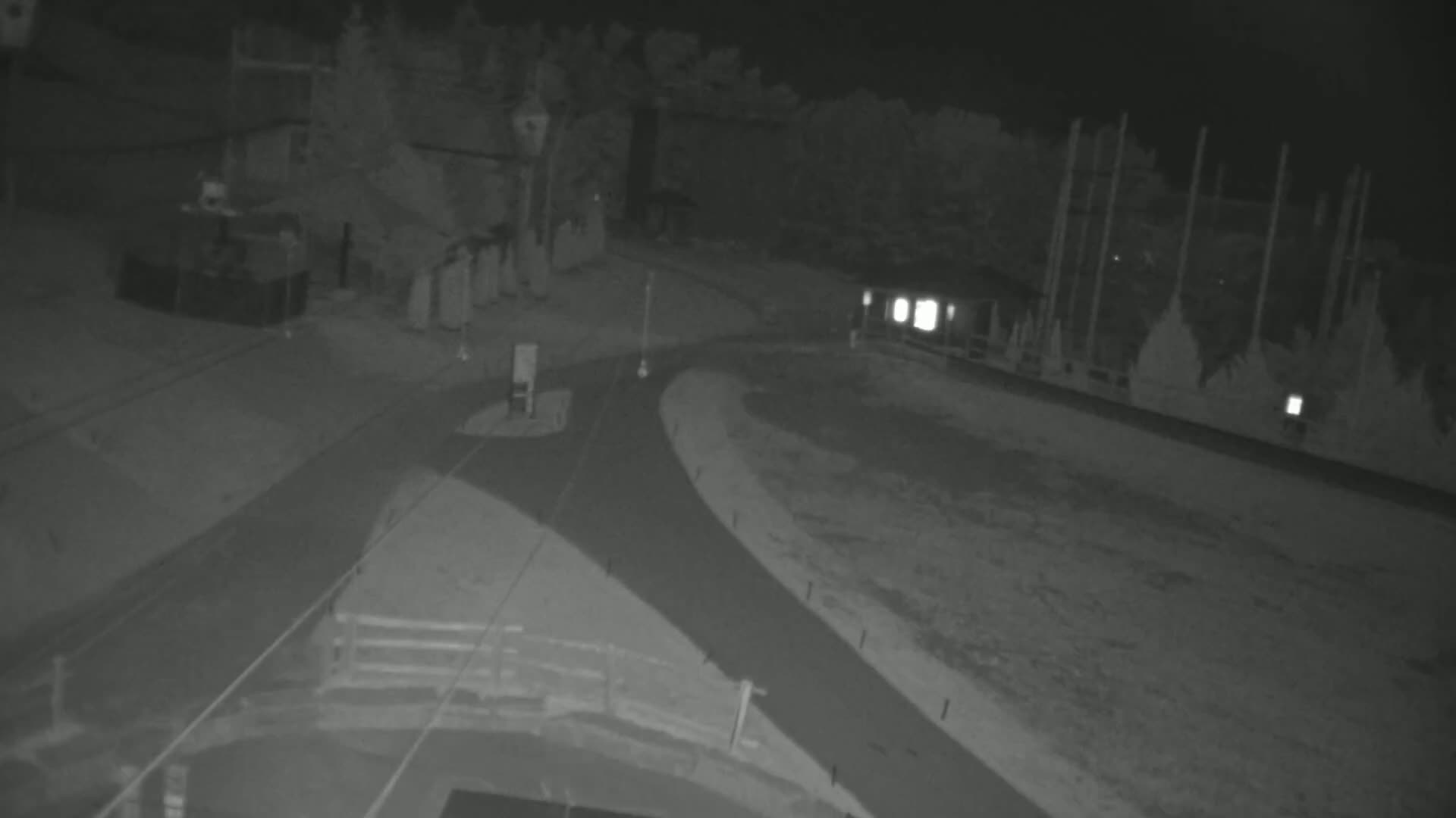 A nighttime, low-light image shows a paved roadway curving through a mostly barren, outdoor area with some structures and trees visible in the background.