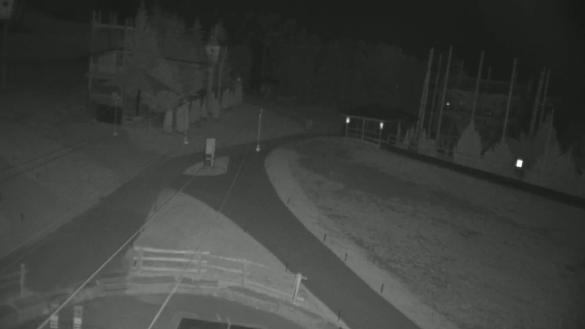 A nighttime, low-light image shows a paved roadway curving through a mostly barren, outdoor area with some structures and trees visible in the background.
