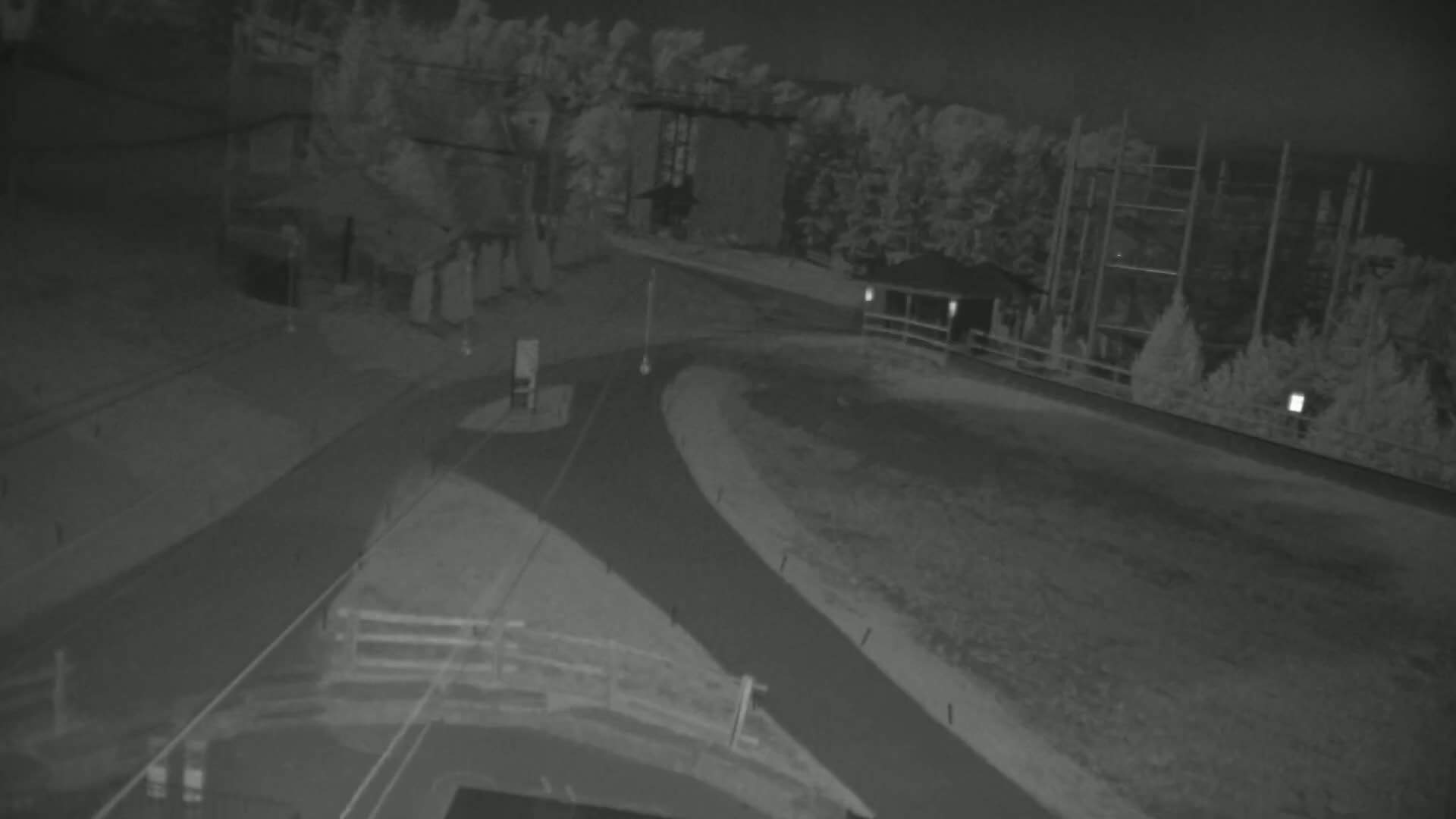 A nighttime, infrared view shows a paved road curving through a sparsely vegetated area with some structures and tall metal supports in the background.