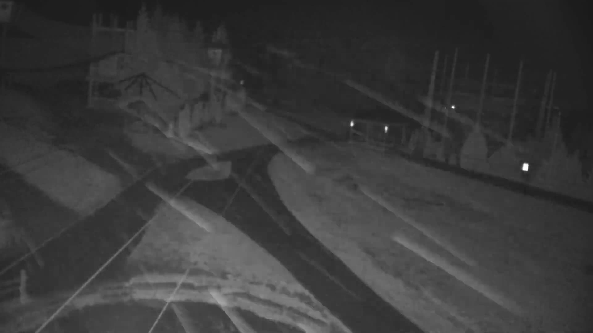 A monochromatic night vision image displays a snow-covered outdoor landscape, possibly a ski resort, featuring buildings, trails, and tall structures, overlaid with bright, ghost-like streaks under cold, clear conditions.
