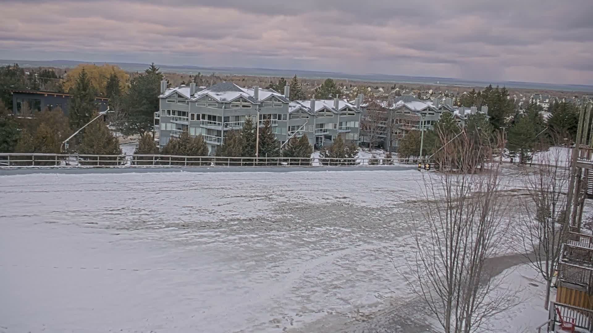 A snow-covered field and fence lead to a complex of multi-story buildings and numerous trees, all under a dark, overcast winter sky with distant water visible on the horizon.