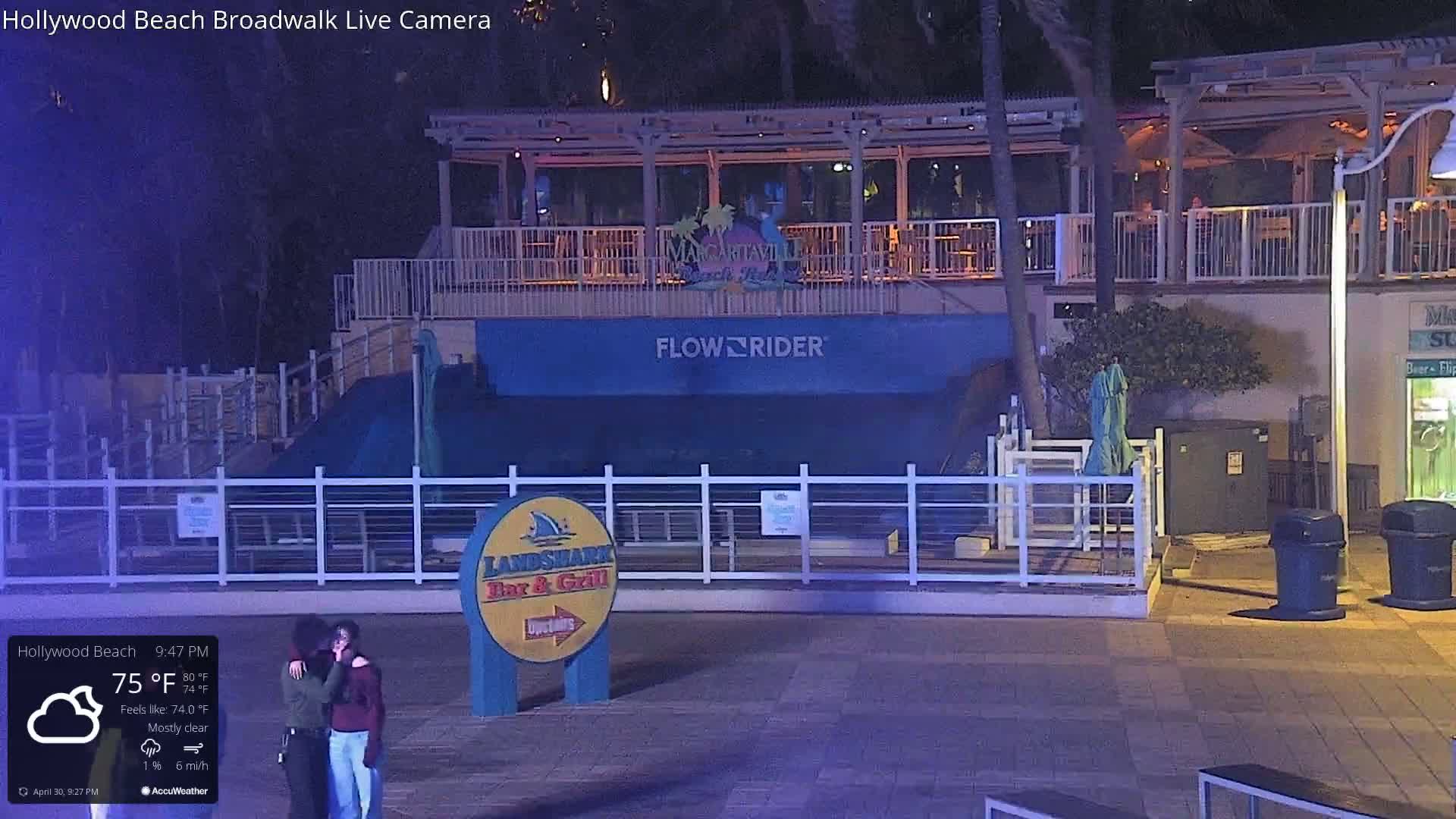 Under a mostly clear night sky, a couple embraces near a Flowrider wave machine and a bar and grill at a boardwalk.