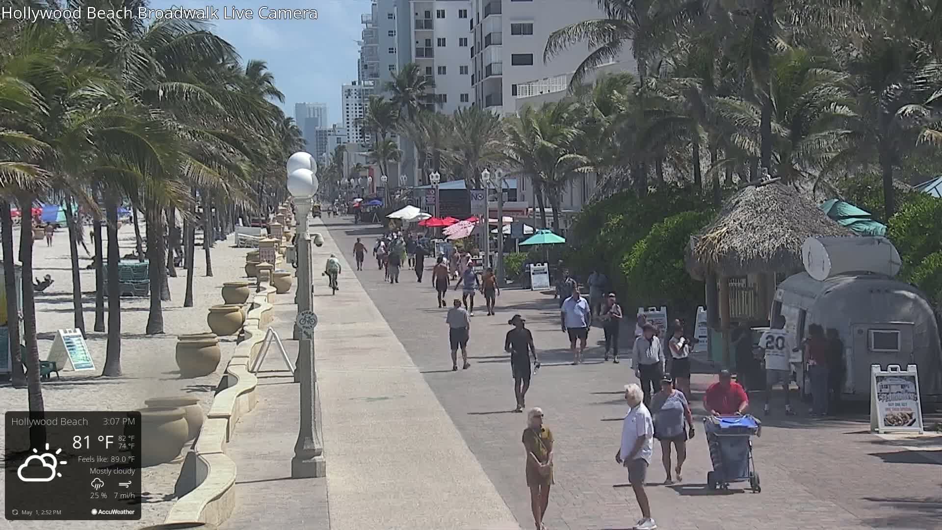 A mostly cloudy day on a beachfront boardwalk shows many people walking along a paved path next to a sandy beach lined with palm trees and beachside businesses.
