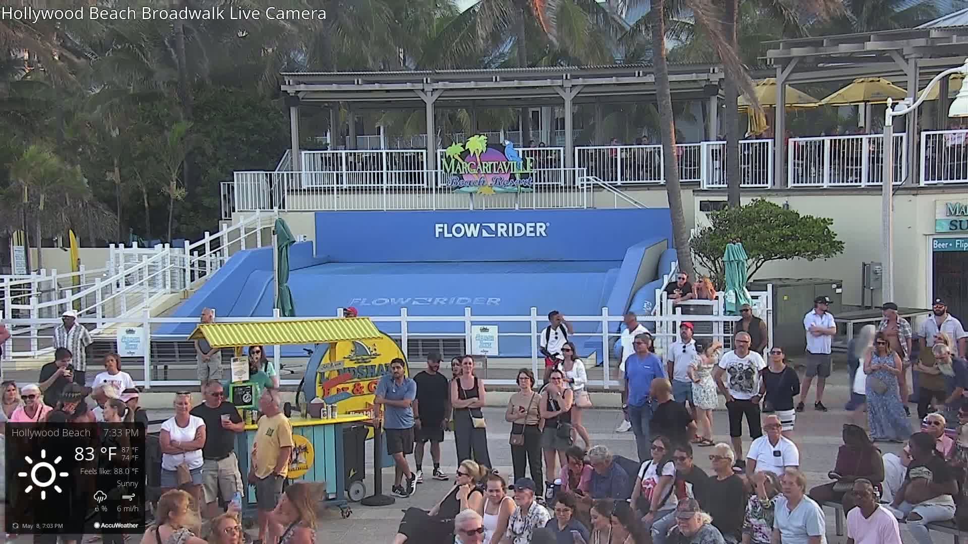 A large crowd gathers around a FlowRider wave machine on a sunny evening.