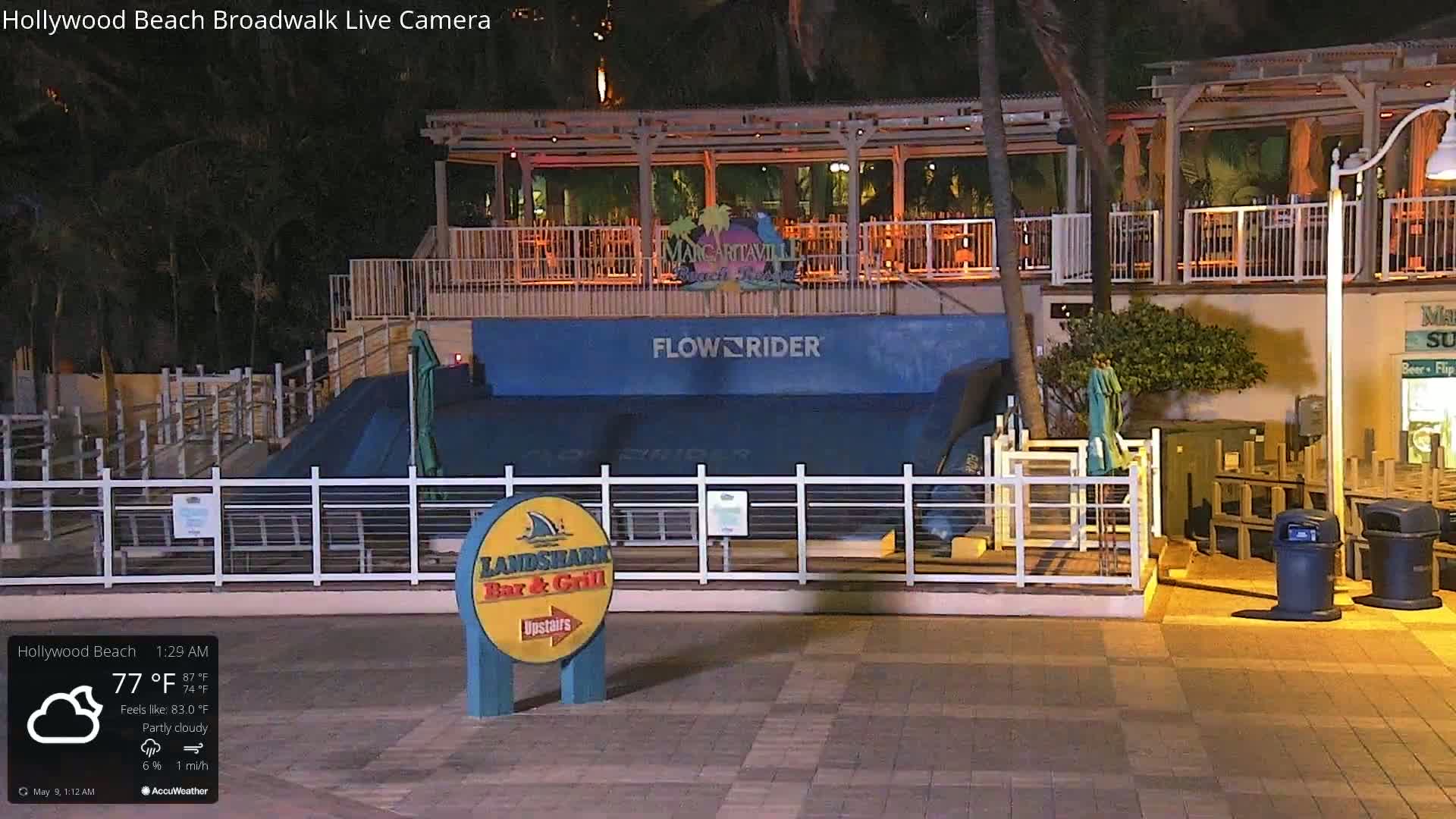 At night under partly cloudy skies, a FlowRider surf simulator sits enclosed by a white fence near a building with outdoor seating.