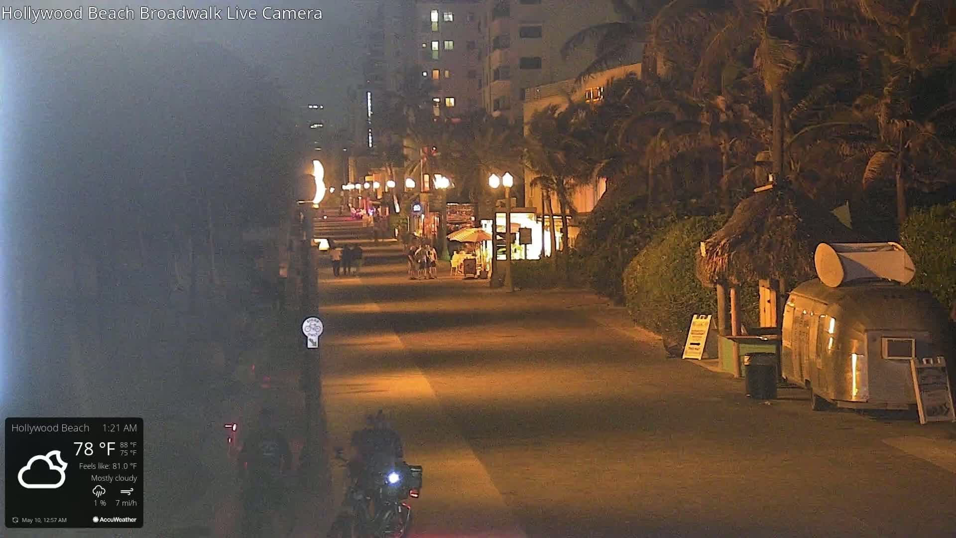 A mostly cloudy night scene shows a mostly empty boardwalk lined with palm trees, buildings, and small shops, with a motorcycle and a few pedestrians visible.