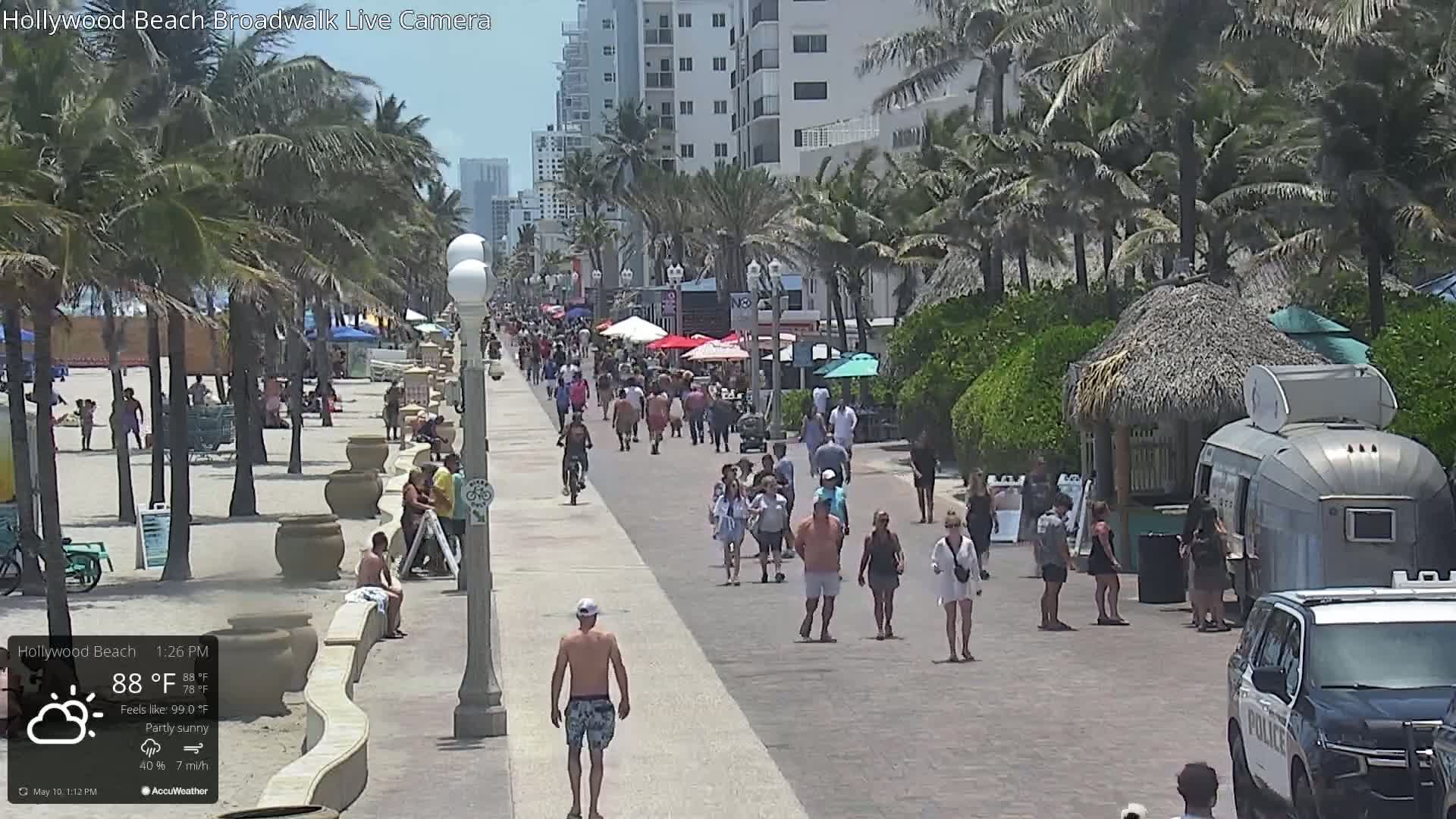 A partly sunny beach boardwalk with many people walking and some sitting on the sand, under a mostly clear sky.