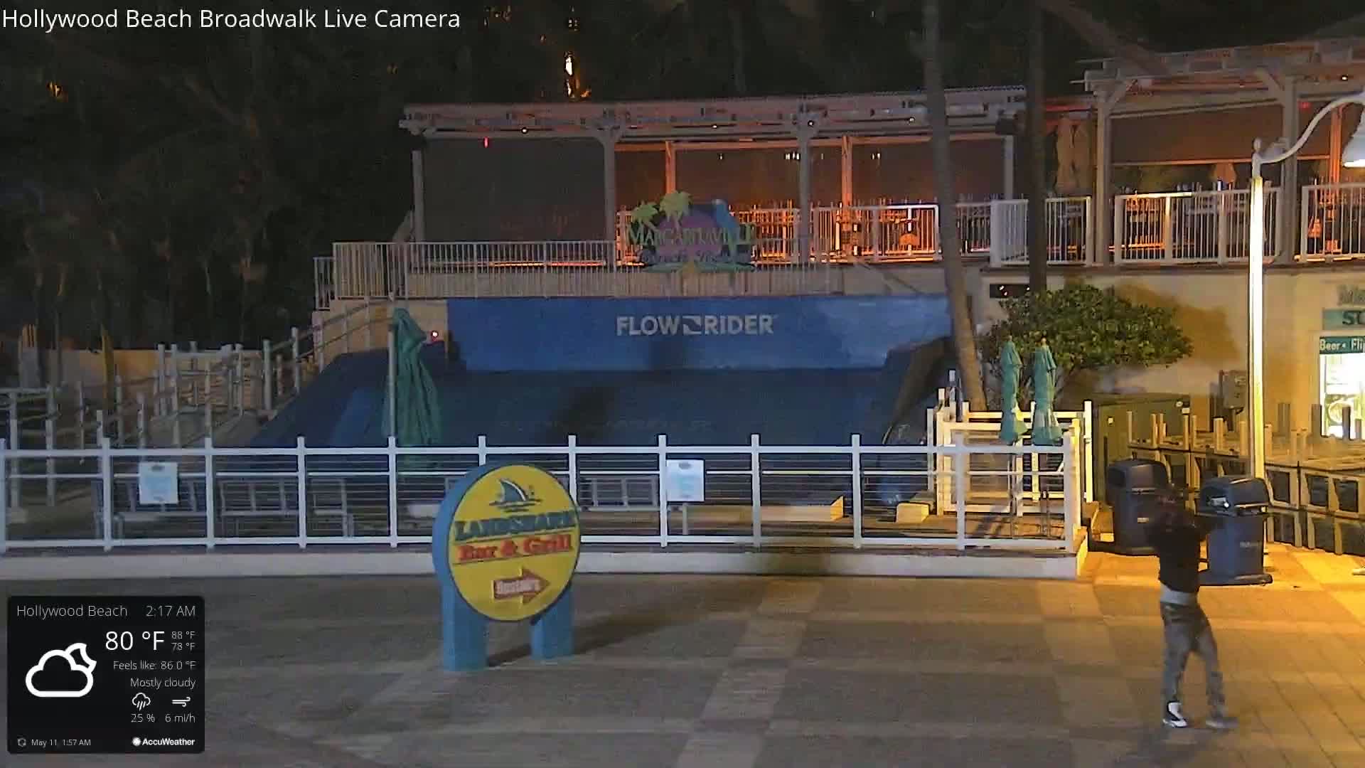 A mostly cloudy night scene shows a person walking near a Flowrider attraction and a bar and grill, with some outdoor seating and trash cans.
