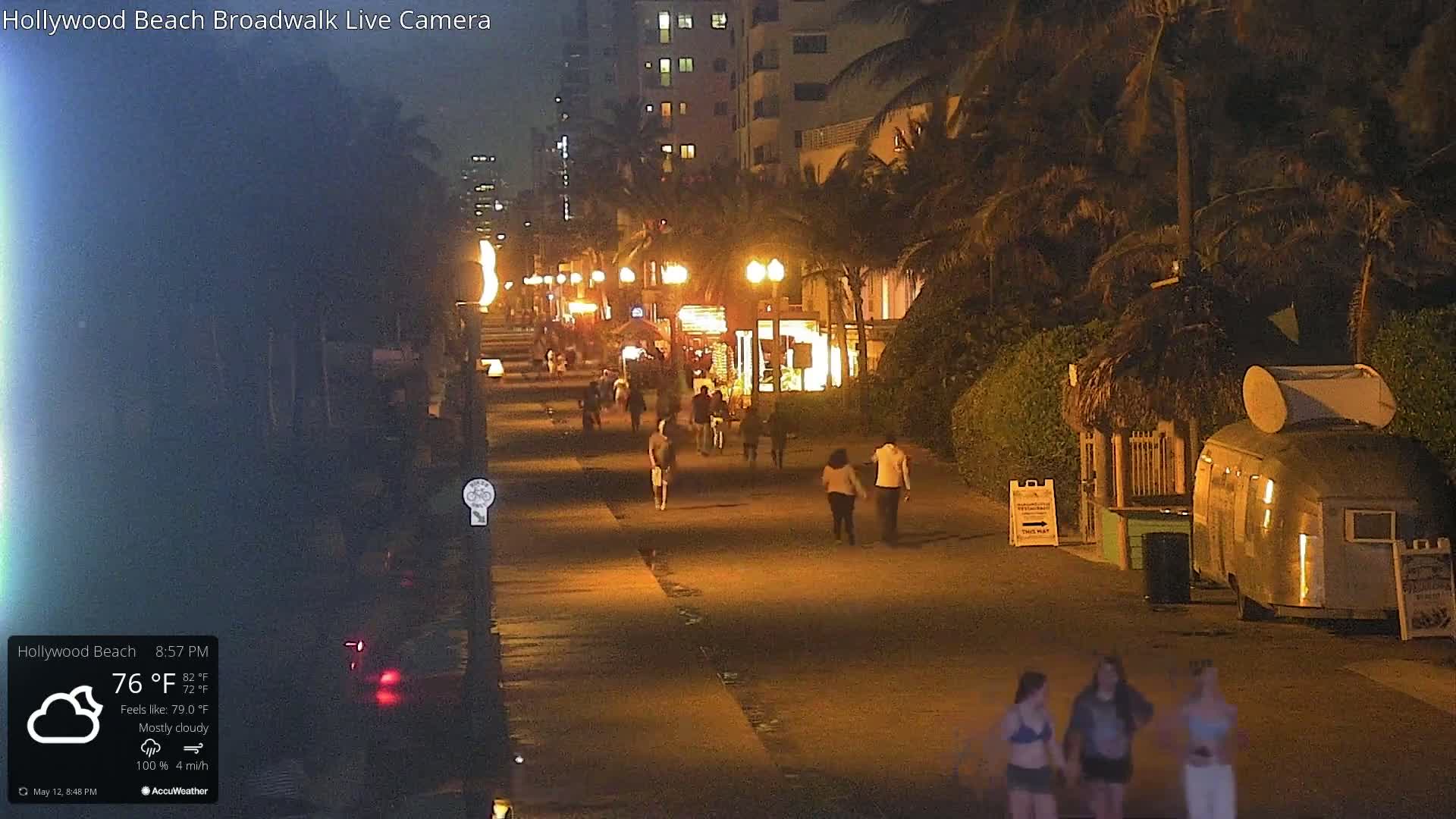 A mostly cloudy night scene shows a boardwalk with people walking under lamplight, palm trees, and buildings lining the street.
