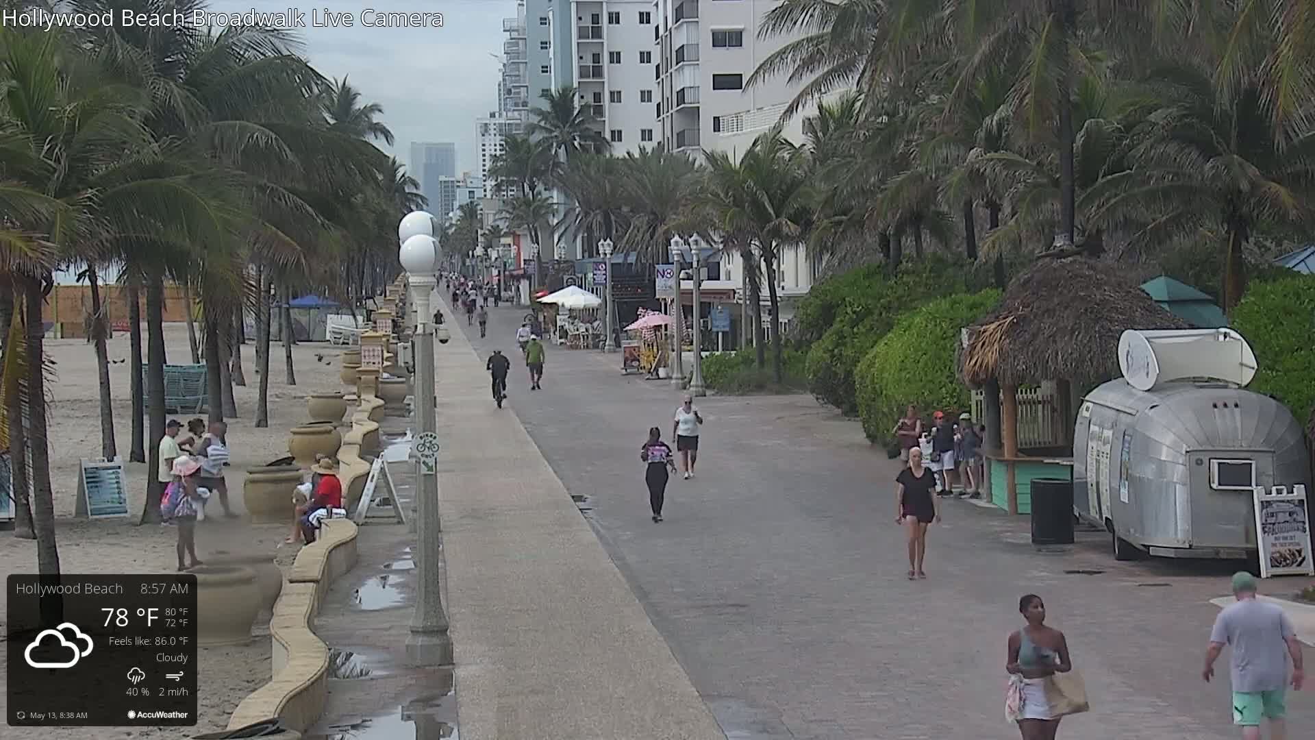A cloudy morning shows a boardwalk with people walking and bicycling along the beach, palm trees, and beachside buildings.