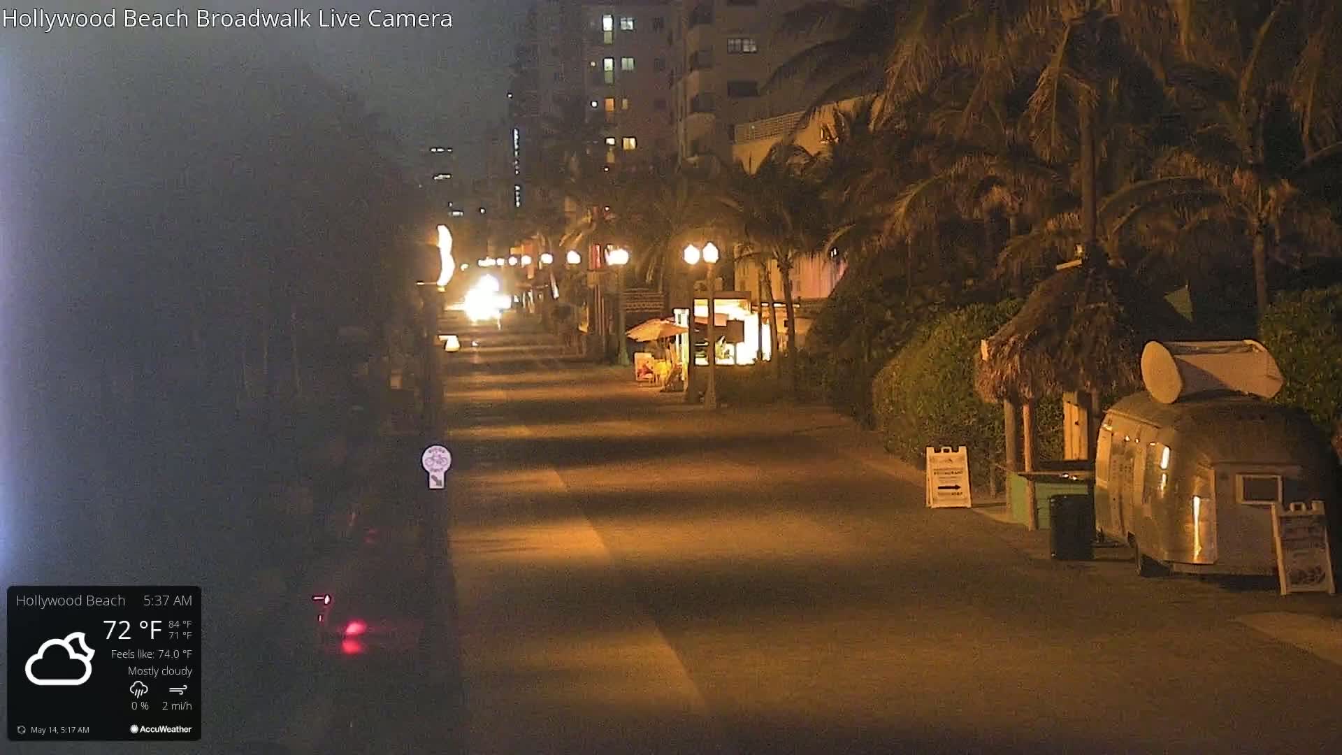 A mostly cloudy night scene shows a mostly empty boardwalk lined with palm trees and a few small businesses, with a food trailer parked nearby.