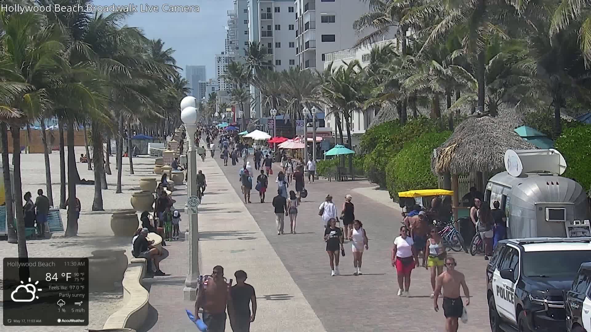 A partly sunny beach boardwalk with many people walking, palm trees, beach umbrellas, and a police car parked nearby.