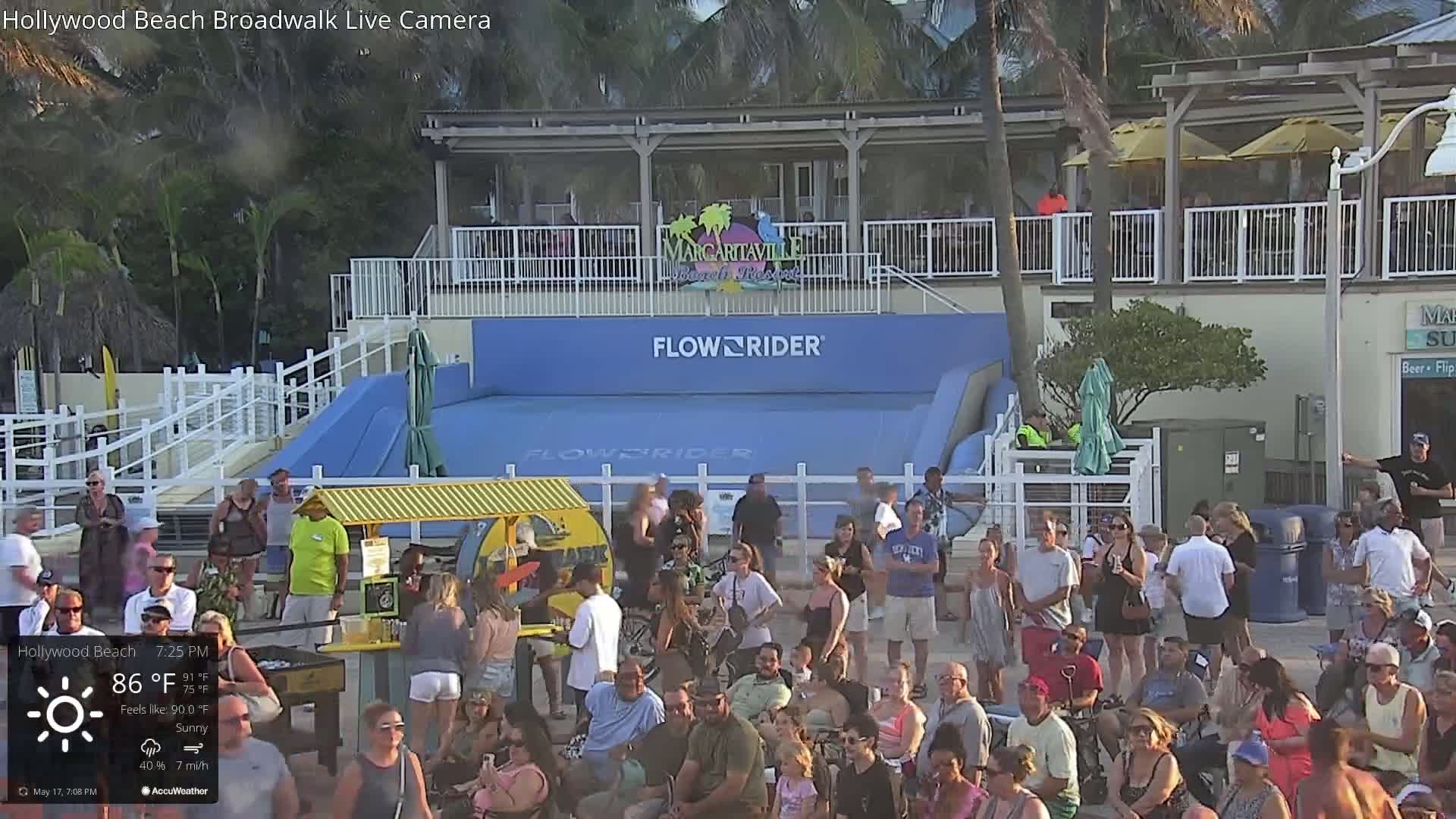 A large crowd of people gathers on a sunny beach near a FlowRider wave machine under a partly cloudy sky.