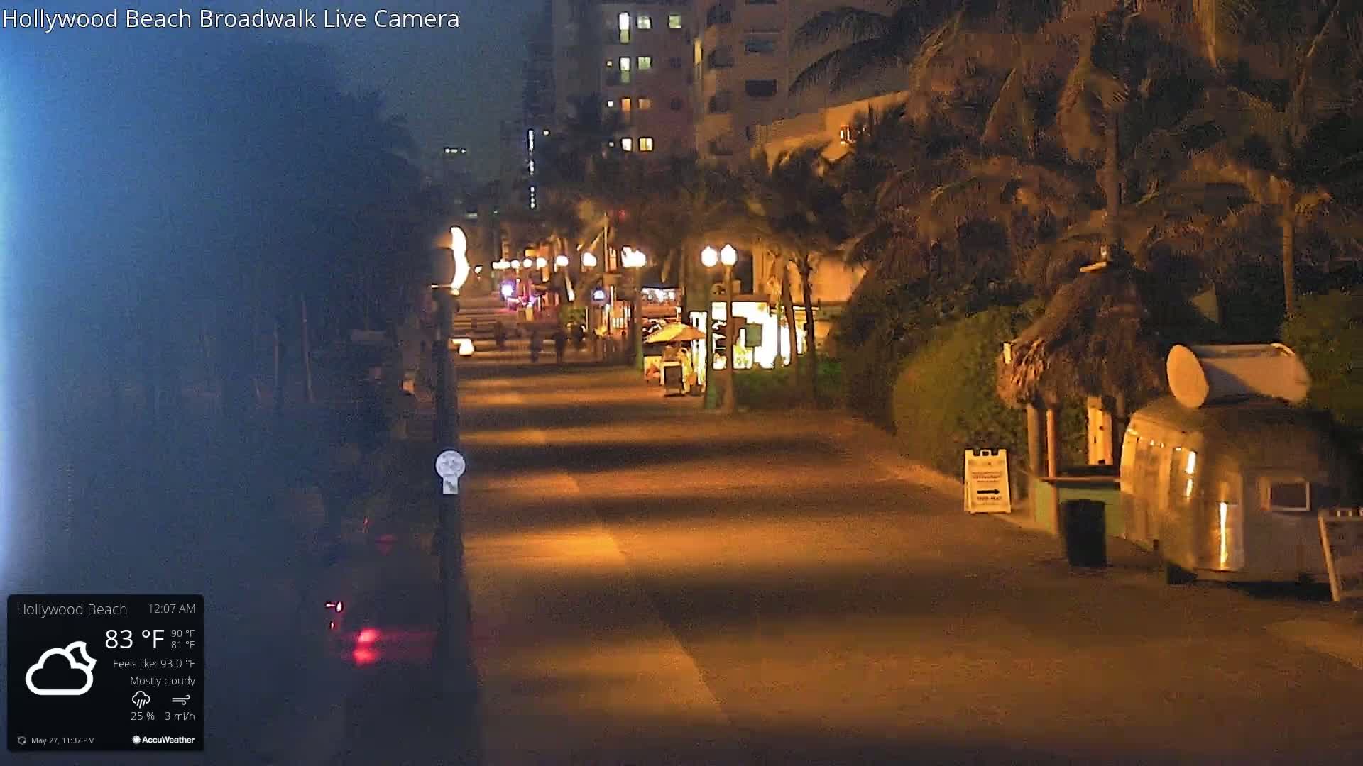 A mostly cloudy night scene shows a mostly empty boardwalk lined with palm trees and buildings, with a few people visible in the distance.
