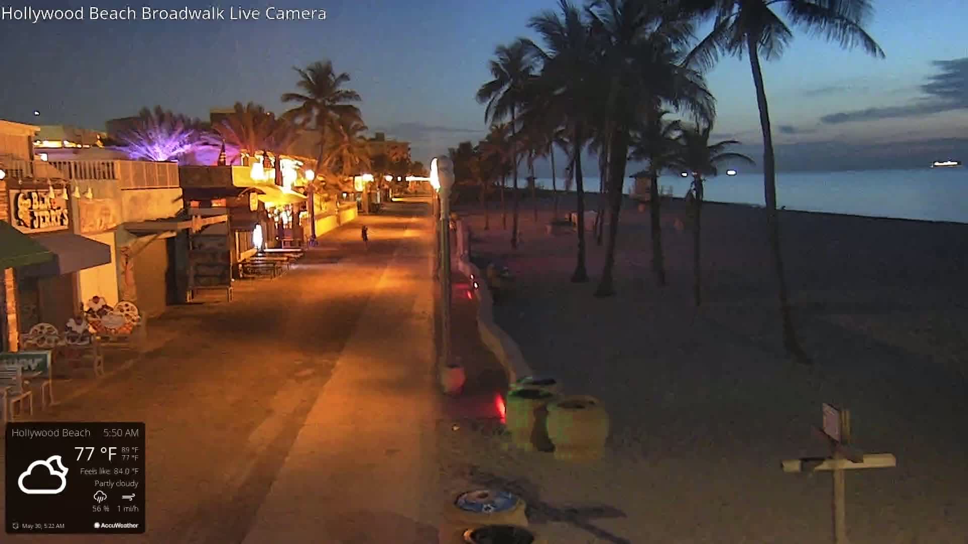 A mostly empty, nighttime boardwalk lined with illuminated shops and palm trees leads to a dark beach under a partly cloudy sky.