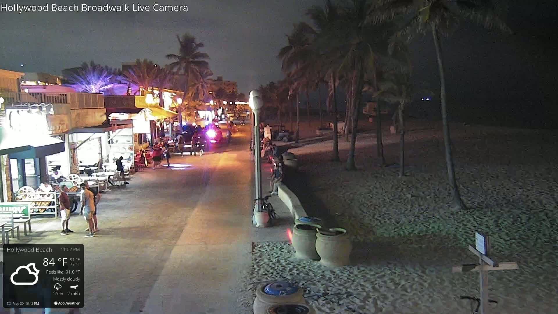 A mostly cloudy night scene shows a boardwalk lined with shops and people, next to a sandy beach under palm trees.