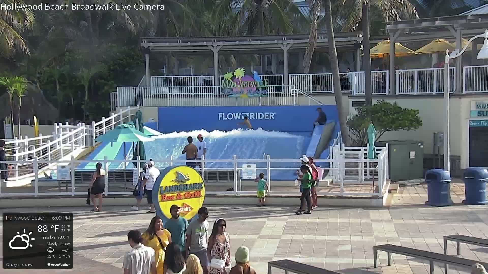 A mostly cloudy outdoor scene shows a Flowrider wave machine with several people around it, along with other people and benches on a paved area.