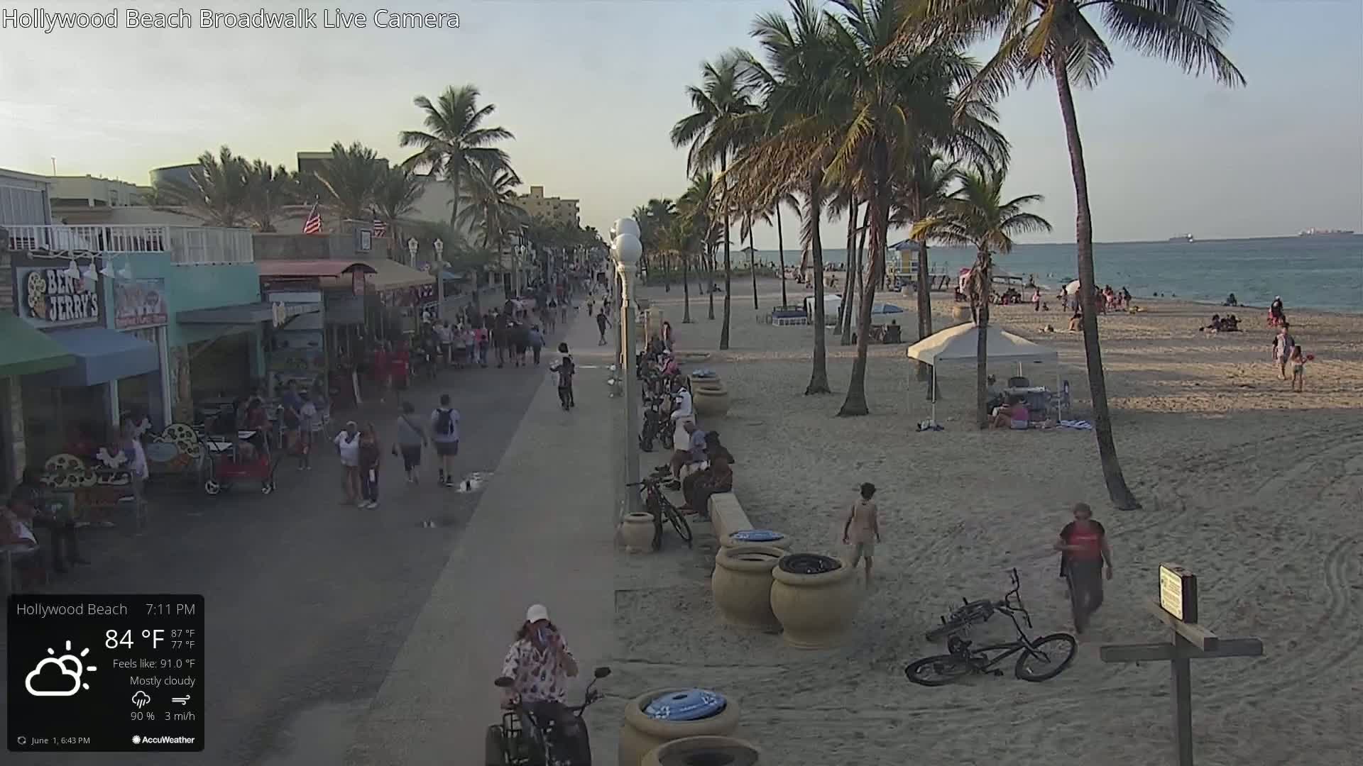 A mostly cloudy evening scene shows a beach boardwalk with people walking, sitting, and biking, palm trees lining the sand, and a calm ocean in the background.
