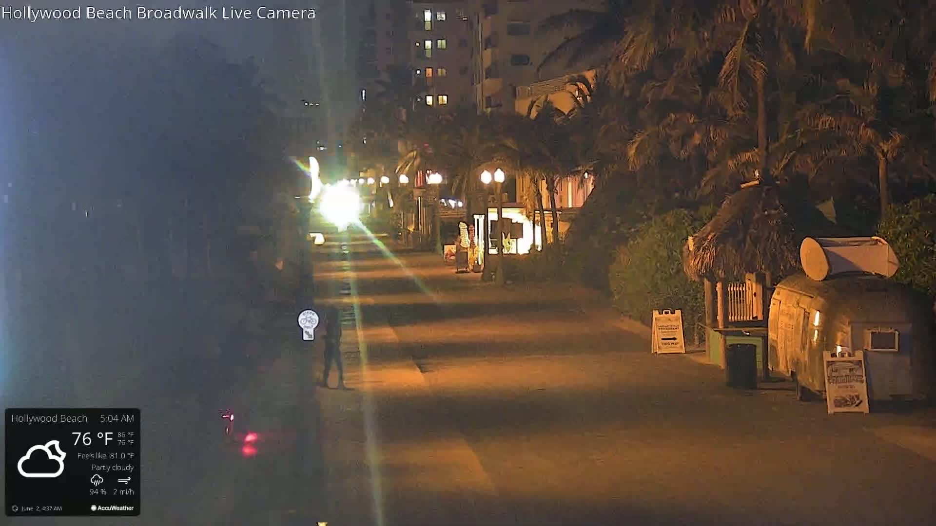 A mostly empty nighttime boardwalk with palm trees and a few small businesses under partly cloudy skies.