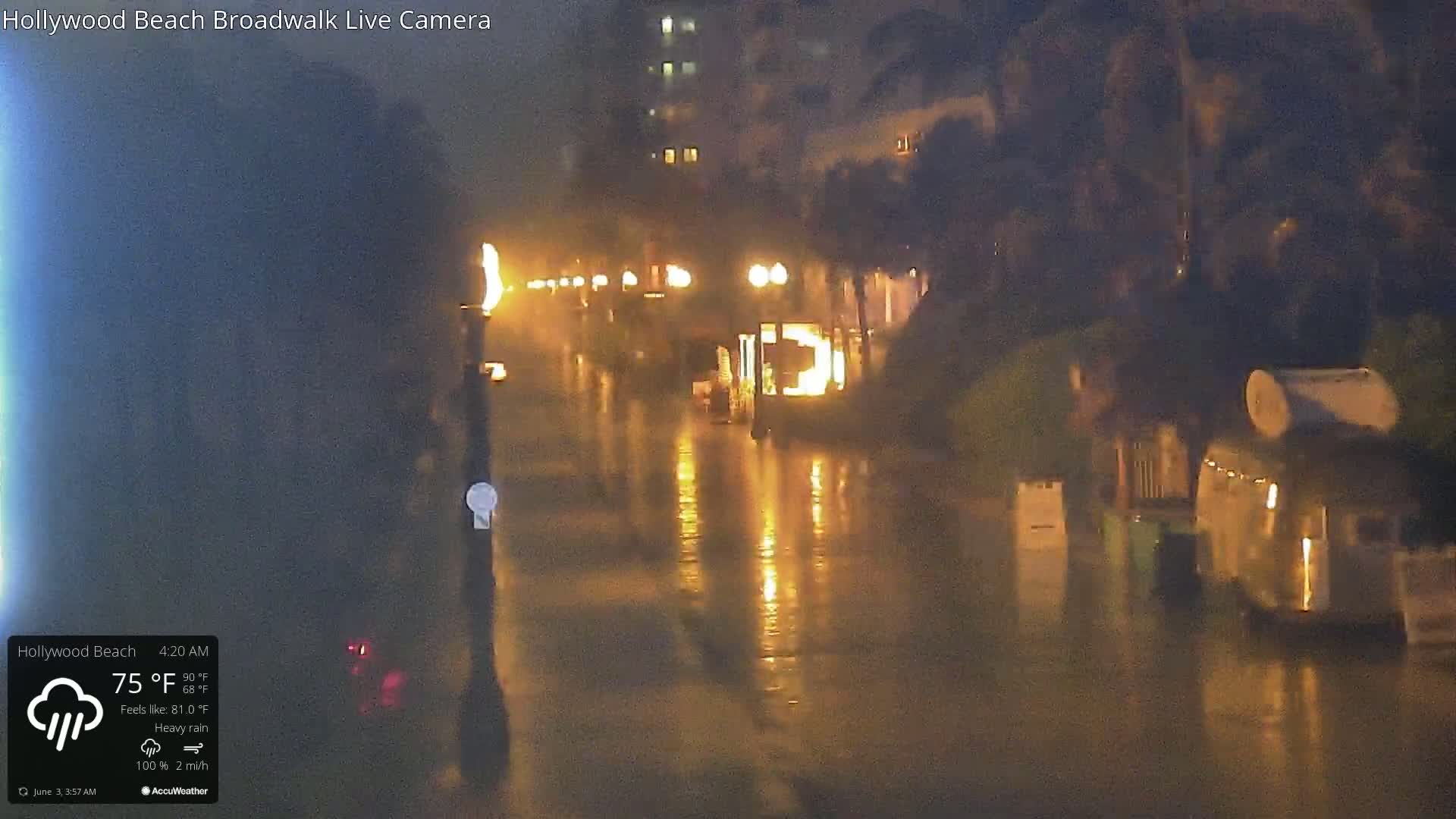 A nighttime street scene shows a flooded boardwalk under heavy rain with illuminated buildings and streetlights reflecting in the water.