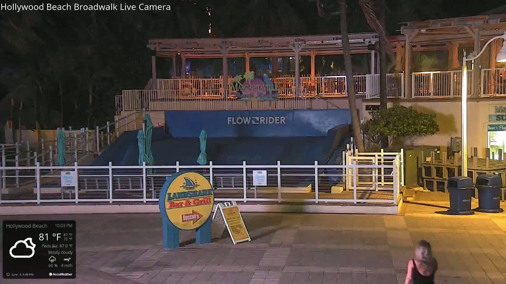 A mostly cloudy nighttime scene shows a FlowRider wave machine at a beachfront establishment, with a person walking in the foreground.