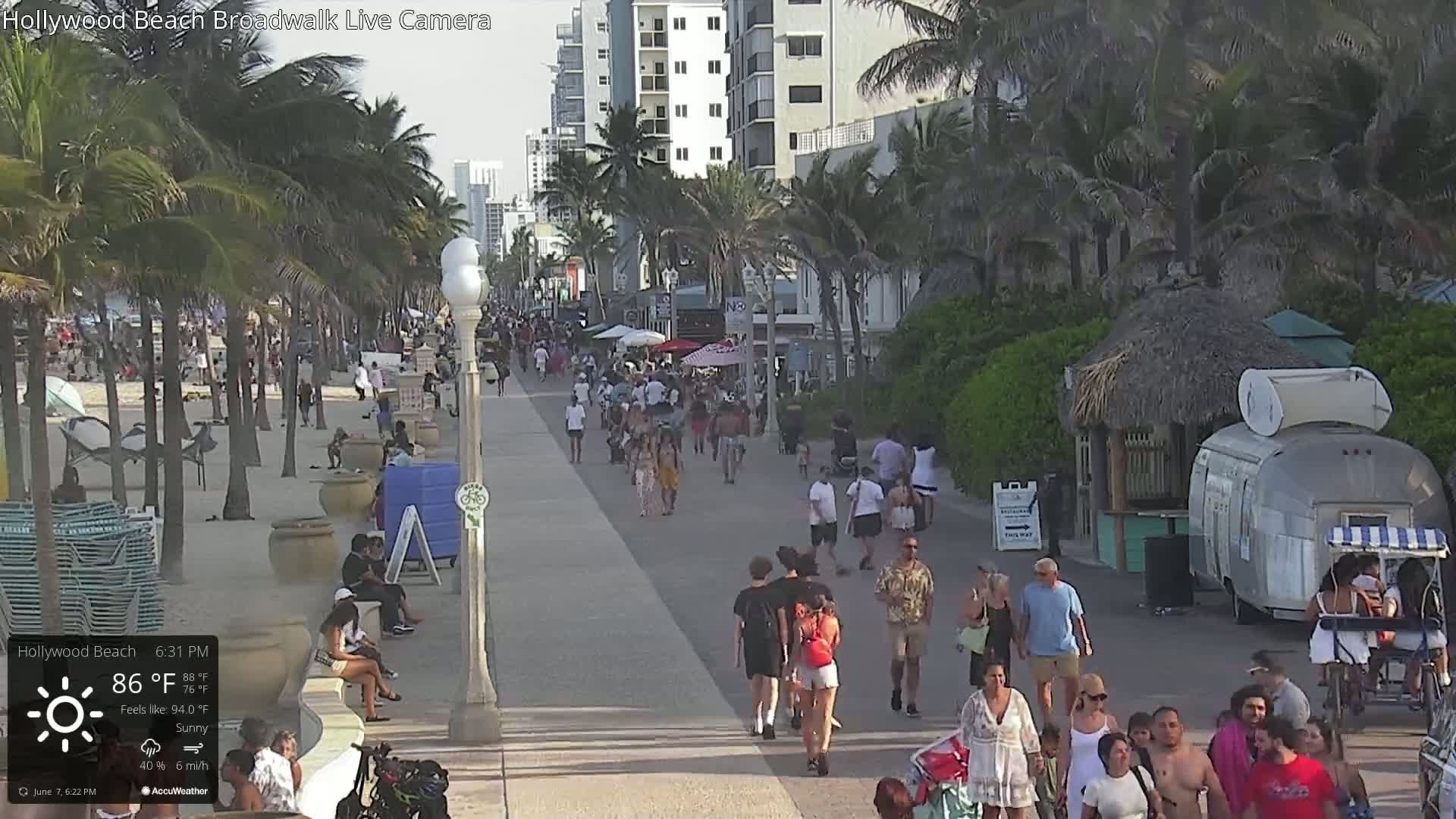 A sunny boardwalk scene shows many people walking along a paved path near a beach with palm trees and beach-front buildings under a partly cloudy sky.