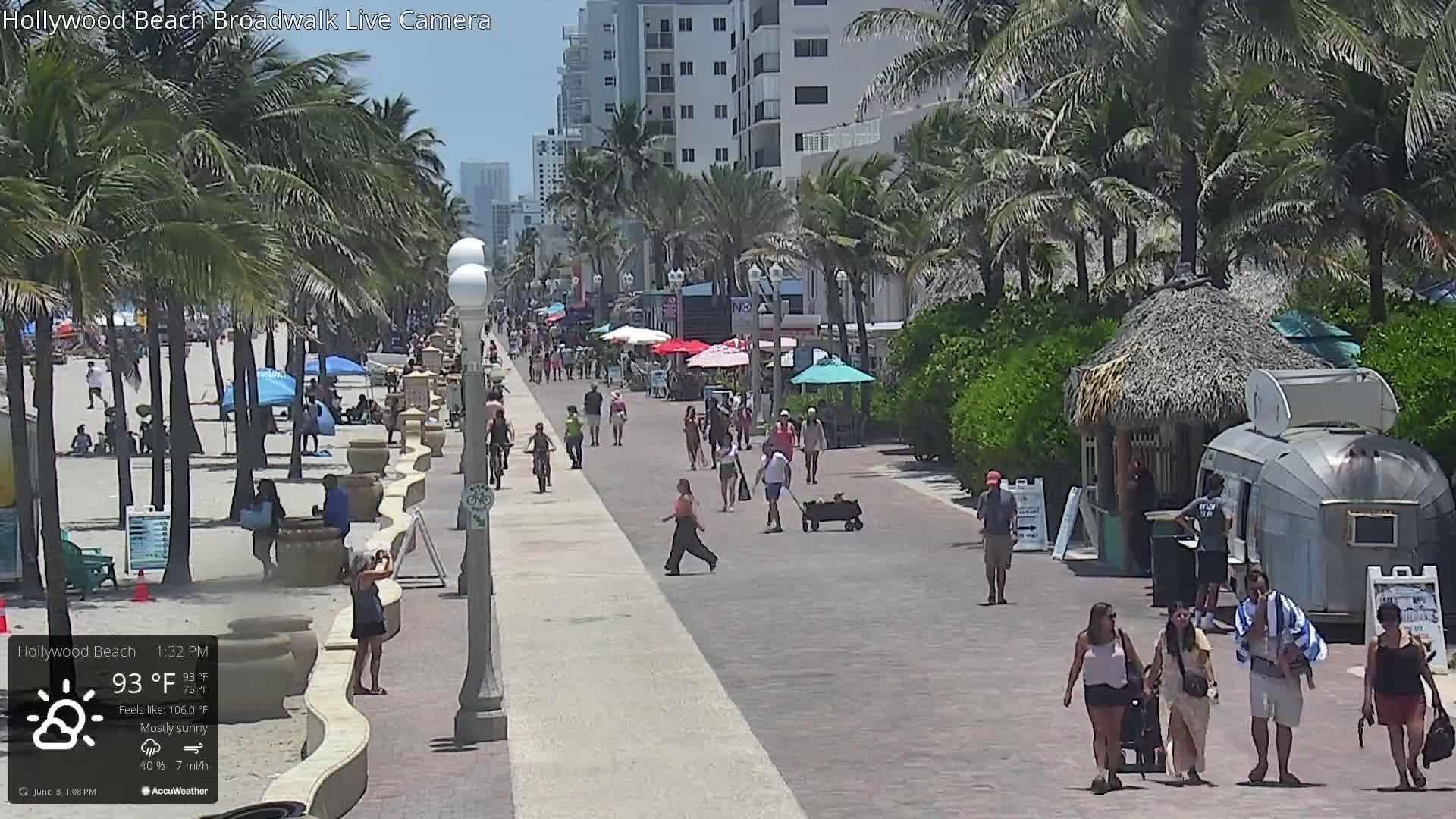 A sunny beach boardwalk with palm trees, people walking, and beach umbrellas on a mostly sunny day.