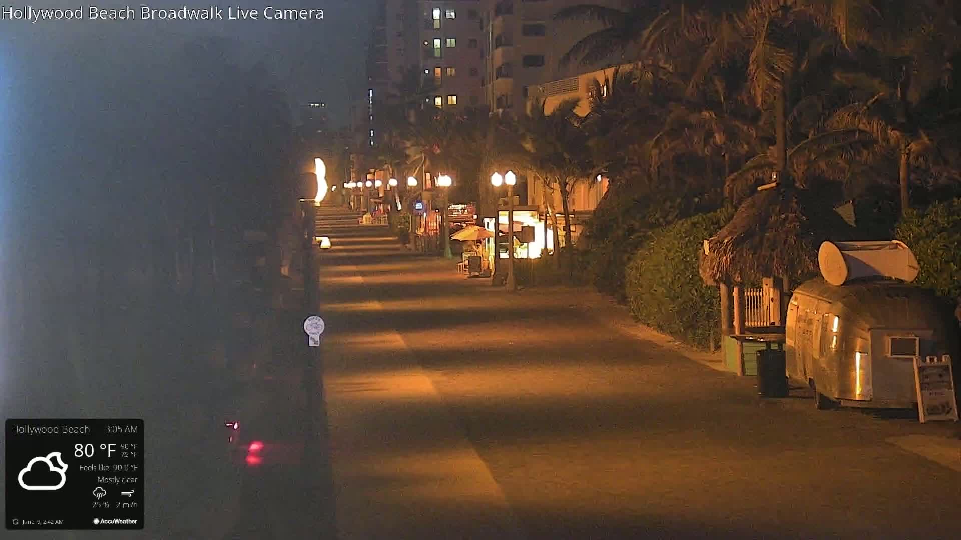 A mostly clear night scene shows a nearly deserted boardwalk lined with palm trees, streetlights, and small businesses, including a food trailer.