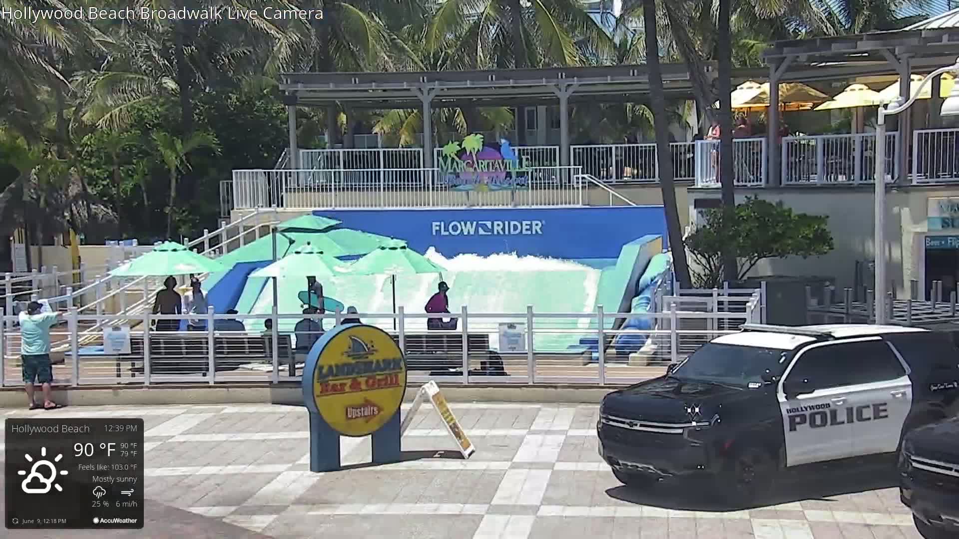 A mostly sunny day shows a FlowRider surf simulator with people using it, beach umbrellas, and a police SUV parked nearby.