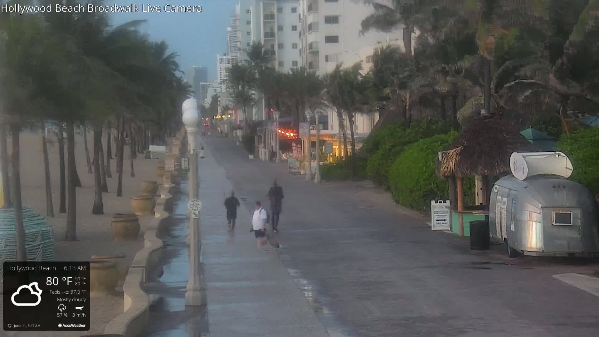 A mostly cloudy morning shows a boardwalk with three people walking, palm trees, and a beachside food stand next to a beach.