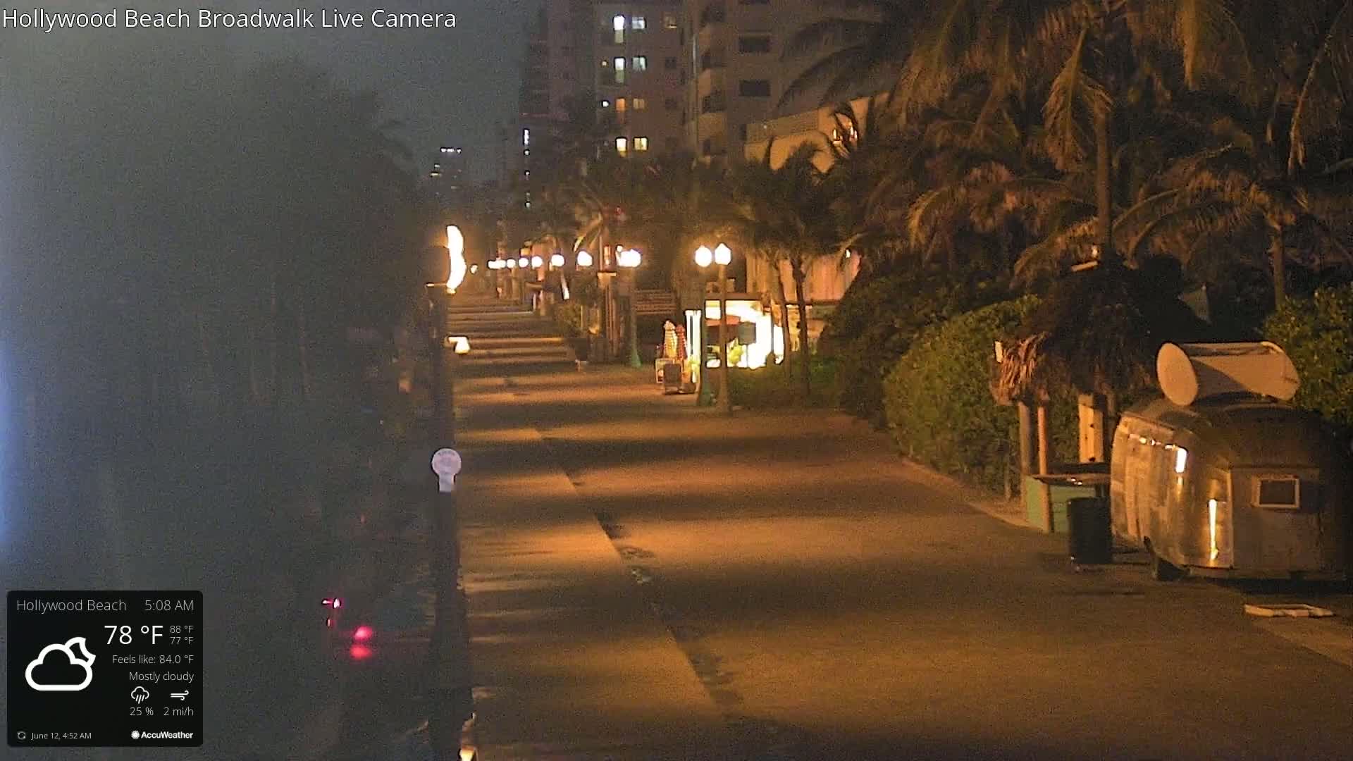 A mostly cloudy nighttime scene shows a paved boardwalk lined with palm trees and small, illuminated shops and kiosks.