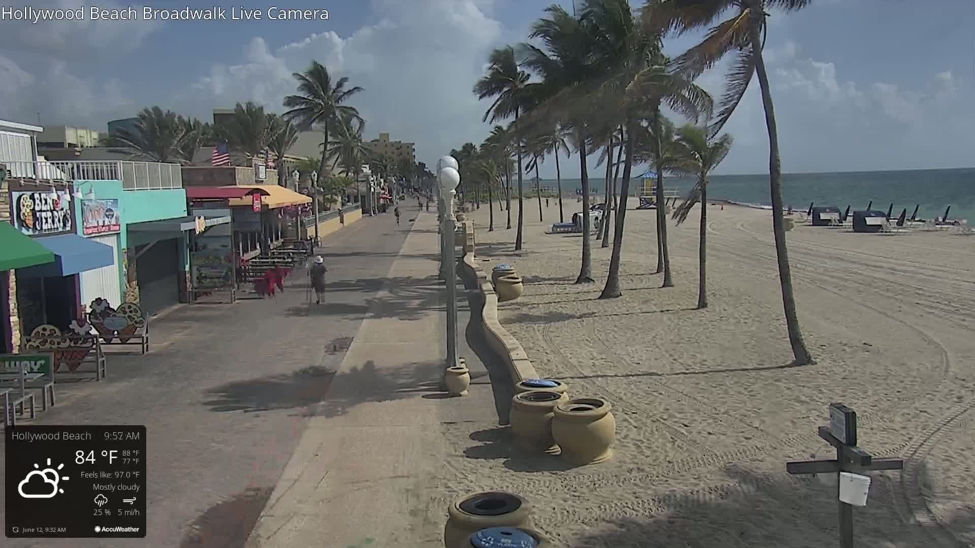 A mostly cloudy day shows a beach boardwalk with palm trees, sand, and several shops and restaurants along the paved walkway.