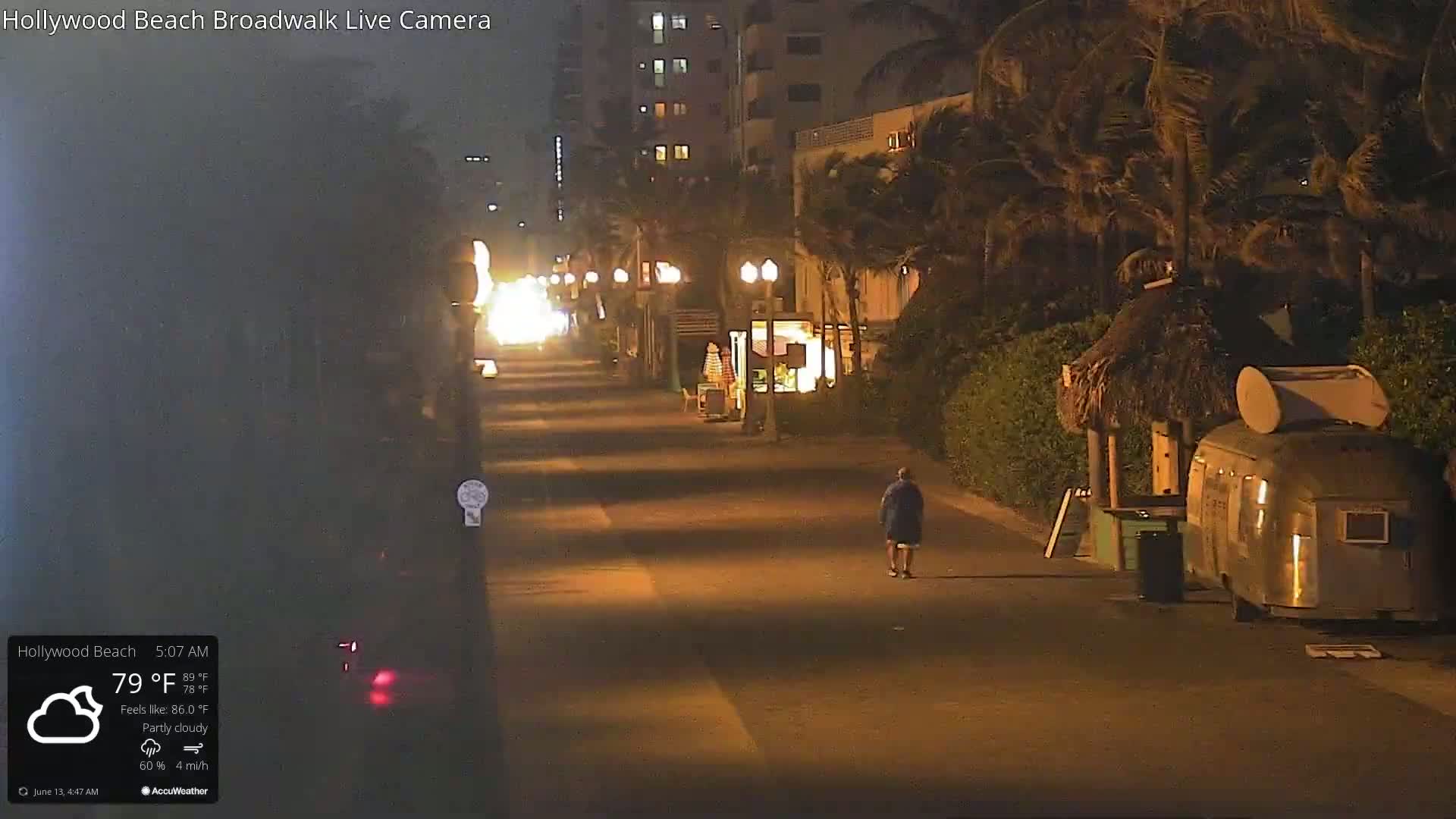 A person walks down a nighttime boardwalk under partly cloudy skies, past buildings, palm trees, and a small hut with a vintage trailer parked nearby.