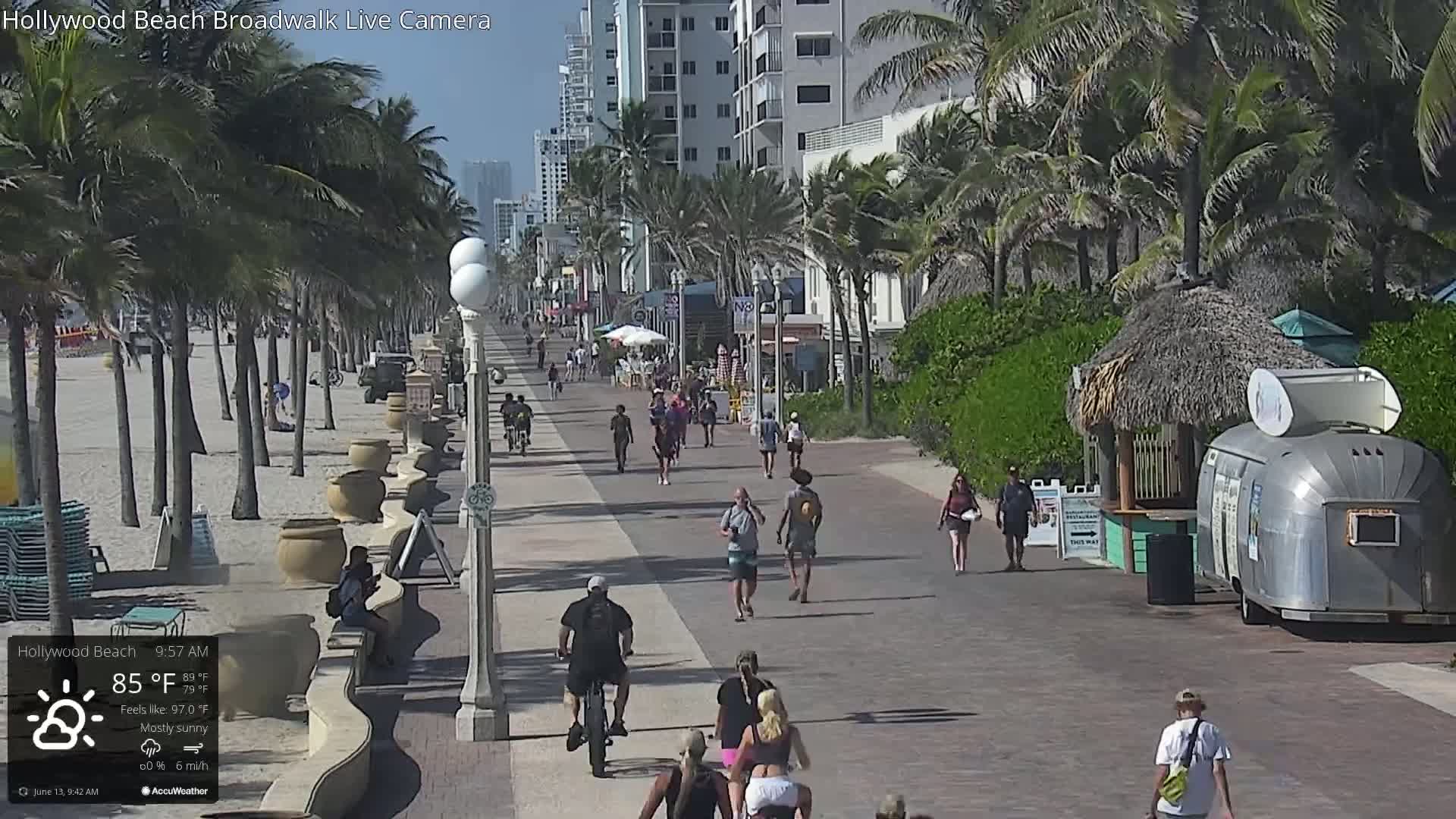 A mostly sunny boardwalk scene shows many people walking and biking along a paved path beside a sandy beach lined with palm trees and beachfront buildings.