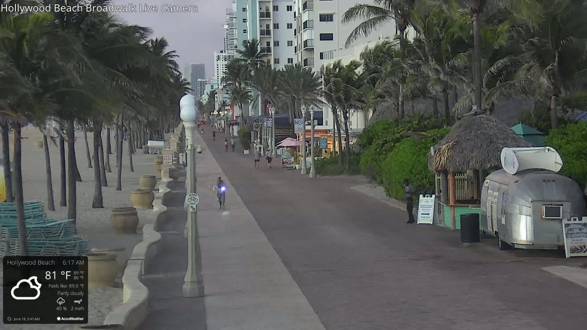 A partly cloudy morning shows a brick boardwalk along a beach lined with palm trees, buildings, and a few pedestrians.