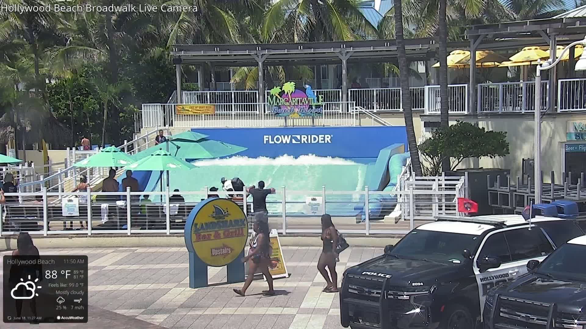 A mostly cloudy day shows a FlowRider wave machine in operation, with people on the boardwalk and two police vehicles parked nearby.