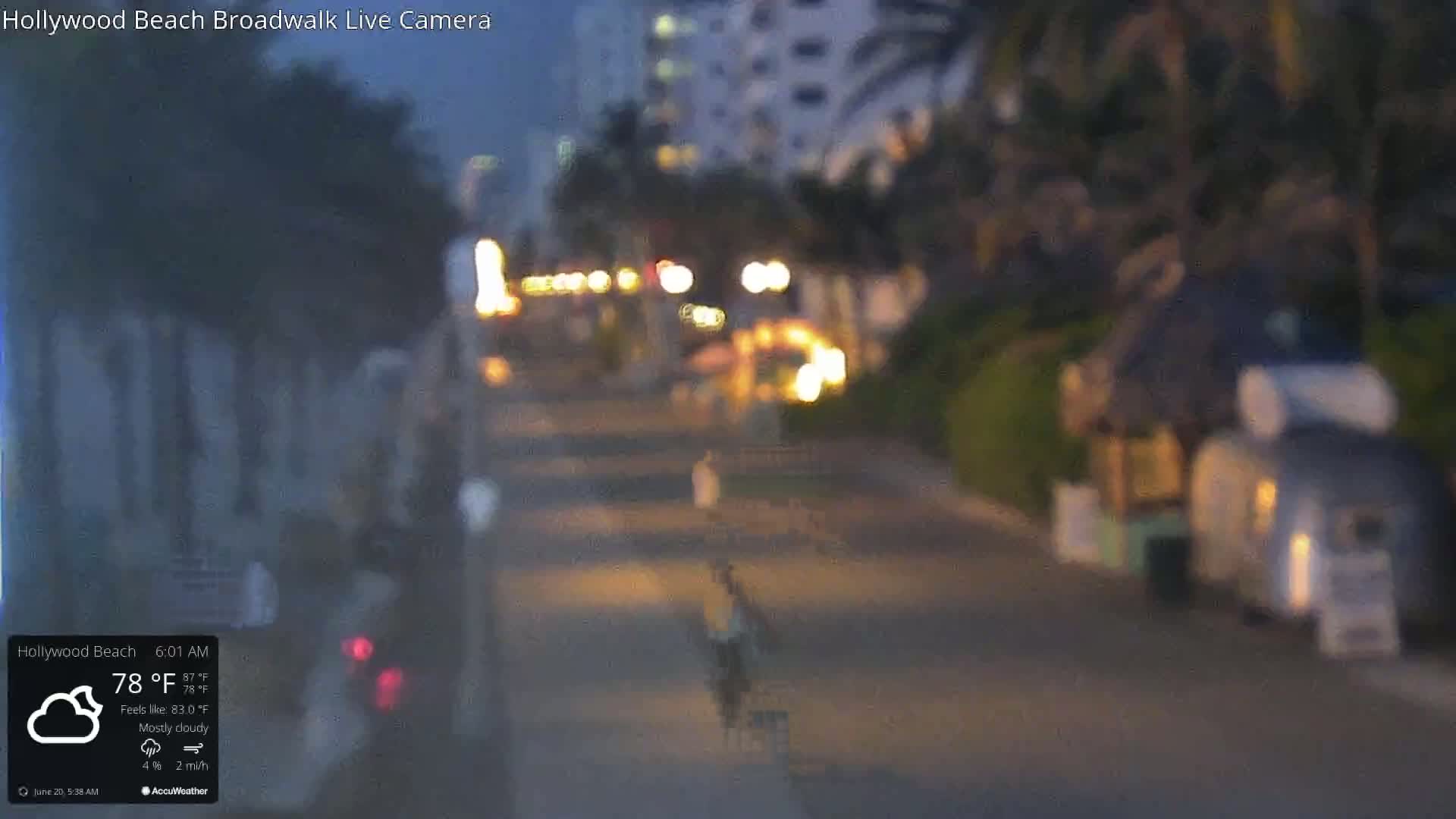A mostly cloudy night scene shows a dimly lit boardwalk with a few people walking, and some buildings and palm trees in the background.