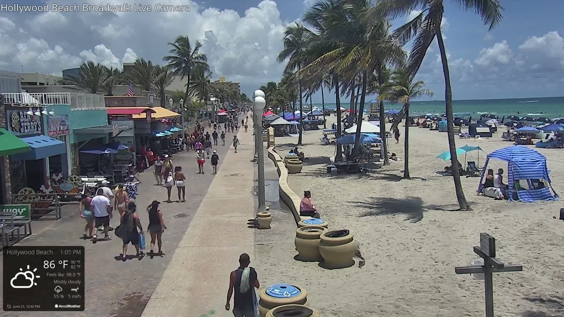 A sunny beach boardwalk scene with mostly cloudy skies shows many people walking along a paved walkway next to a sandy beach lined with palm trees and beach umbrellas.
