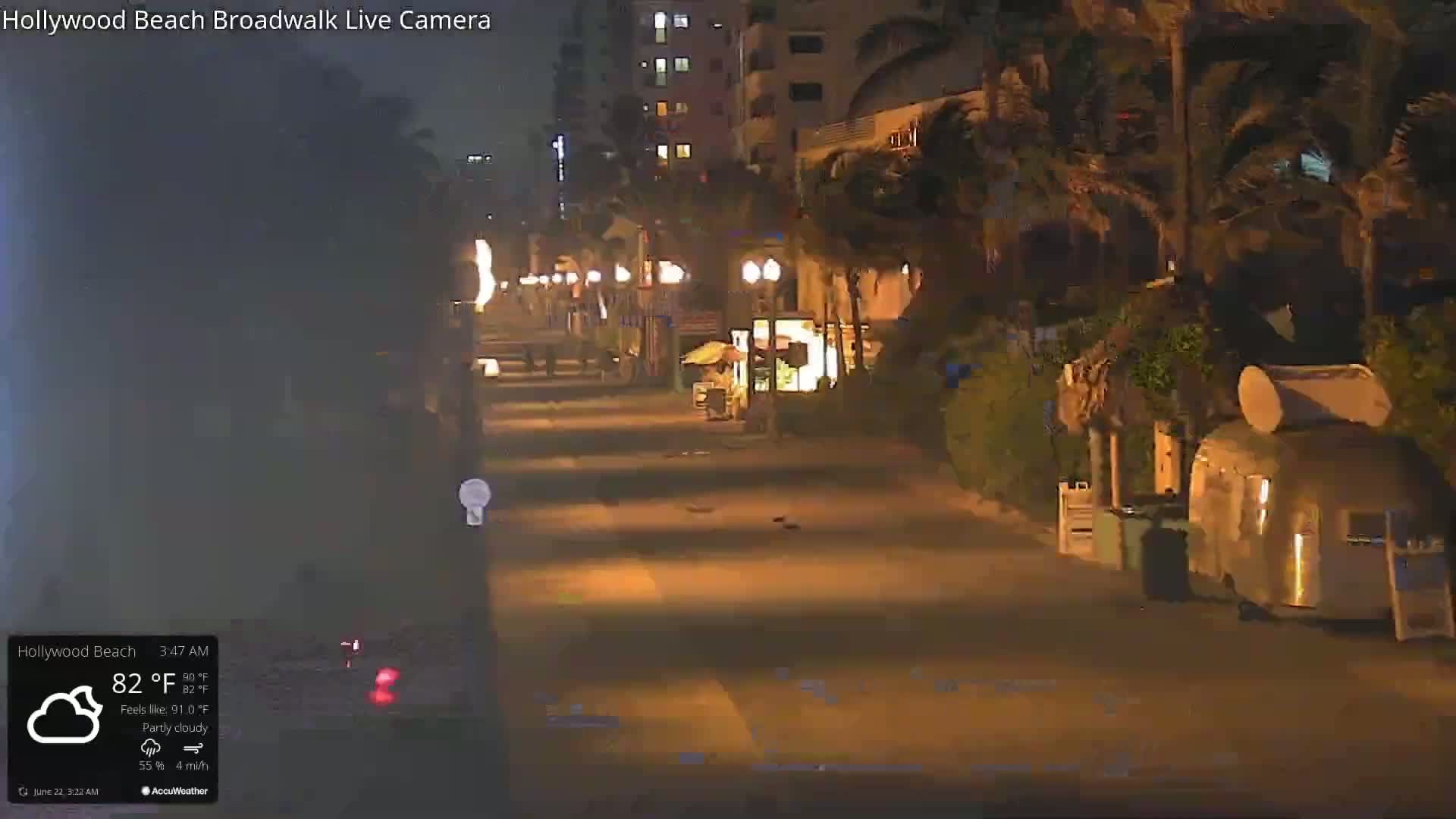 A mostly empty nighttime boardwalk lined with palm trees and a few food stands under partly cloudy skies.
