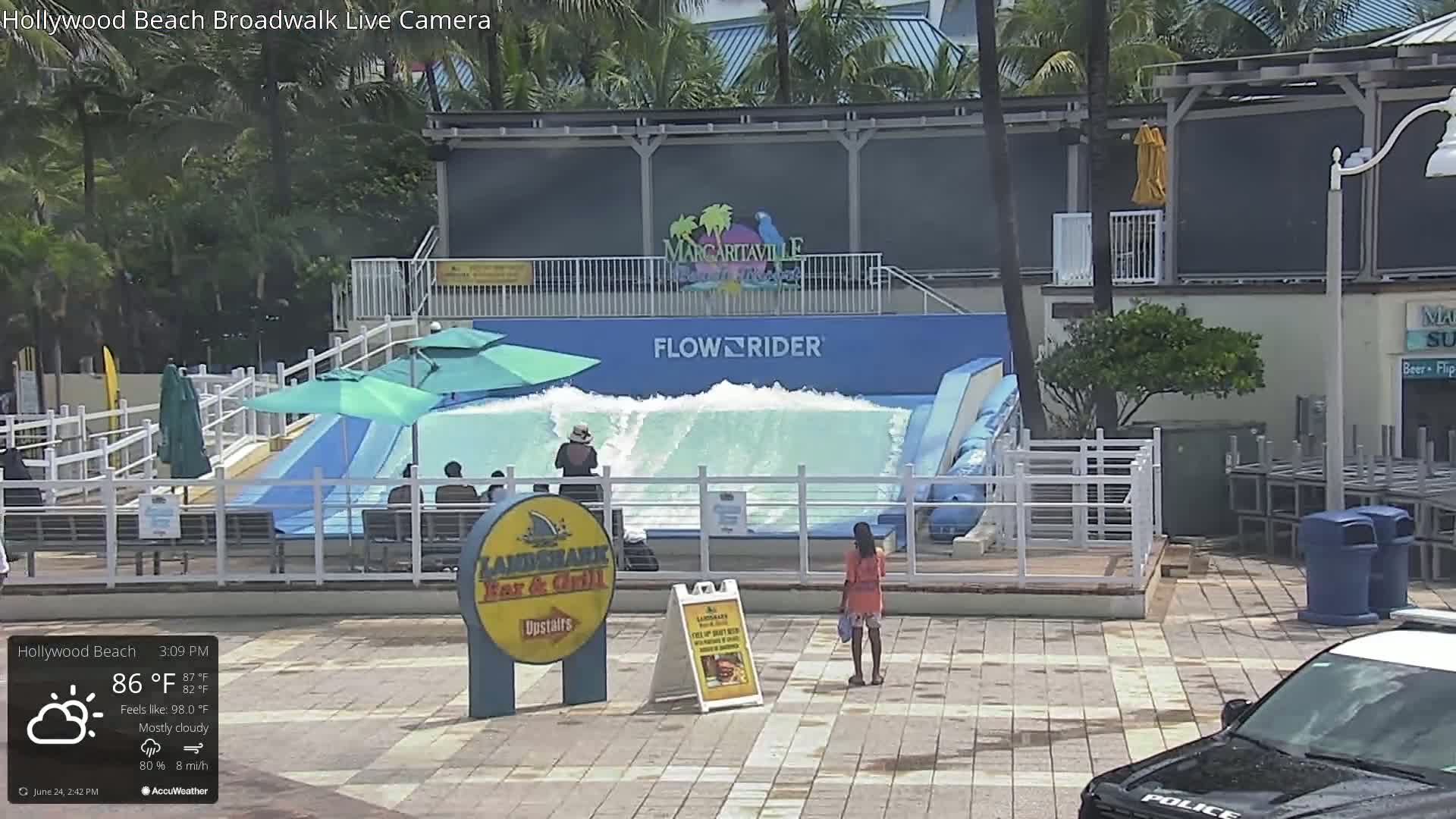 A FlowRider wave machine is operating under mostly cloudy skies, with a few people nearby on a paved area.