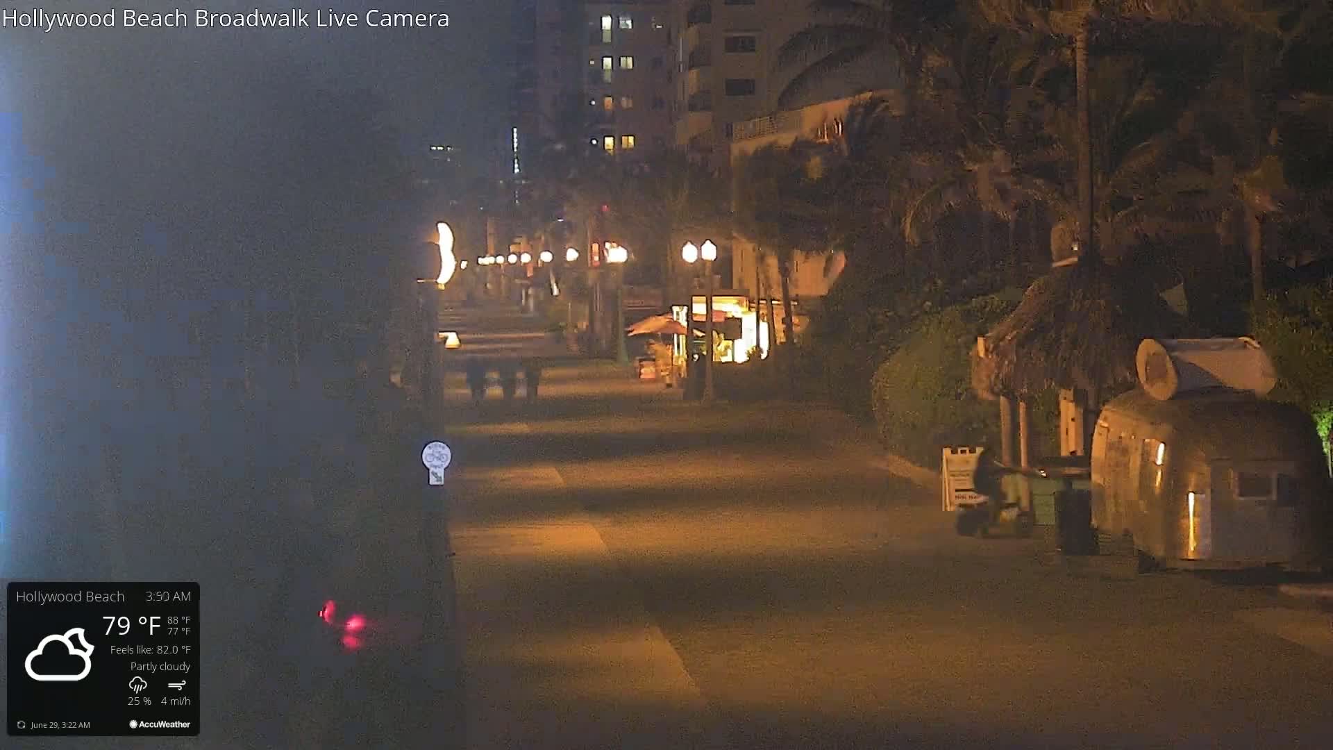 A nighttime, partly cloudy scene shows a boardwalk with a few people walking, palm trees, and small businesses along the sides of the road.