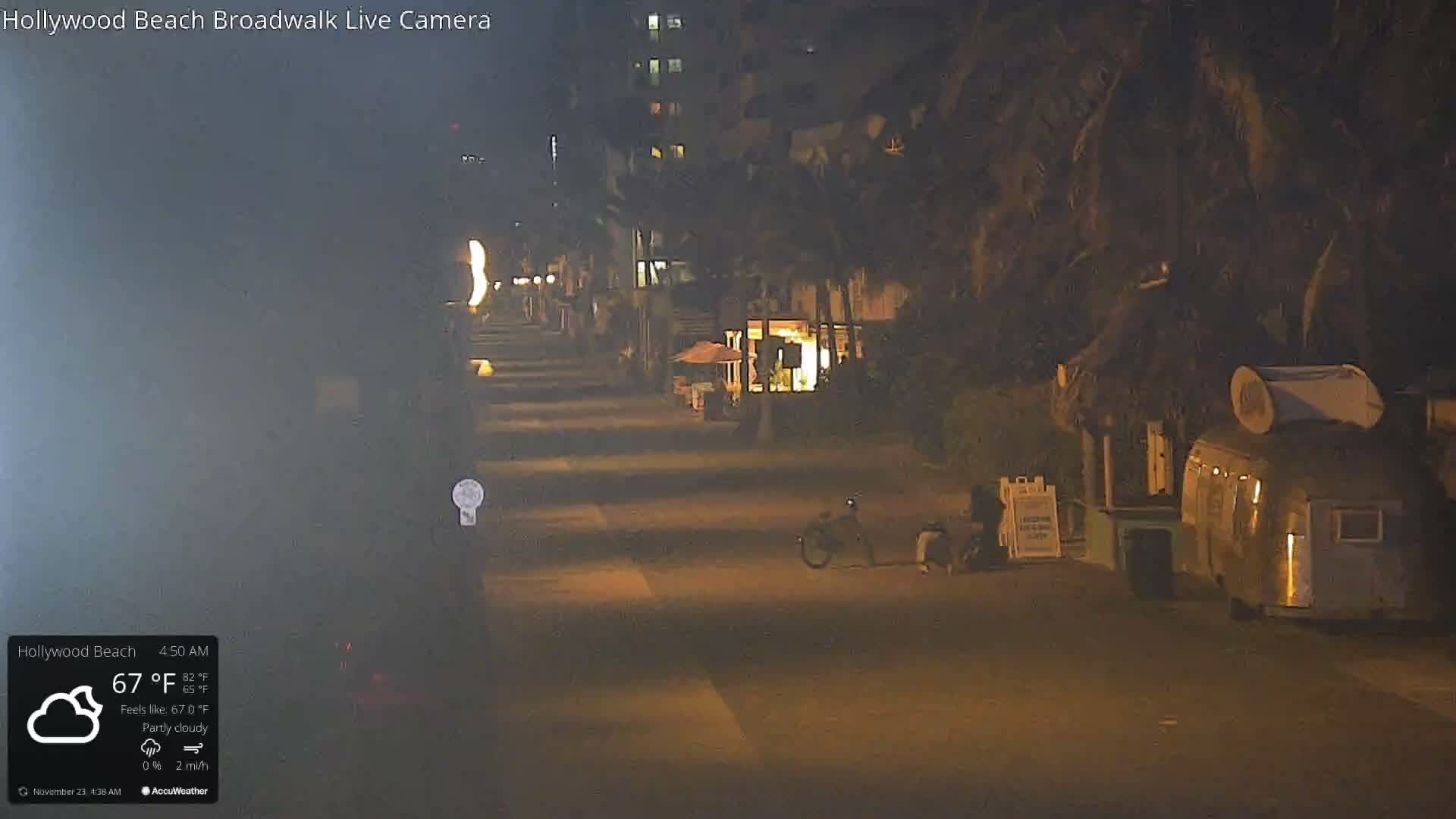 A dimly lit coastal boardwalk scene at early morning shows palm trees, multi-story buildings with illuminated windows in the background, a parked bicycle, and unique vendor stalls, all under partly cloudy skies.