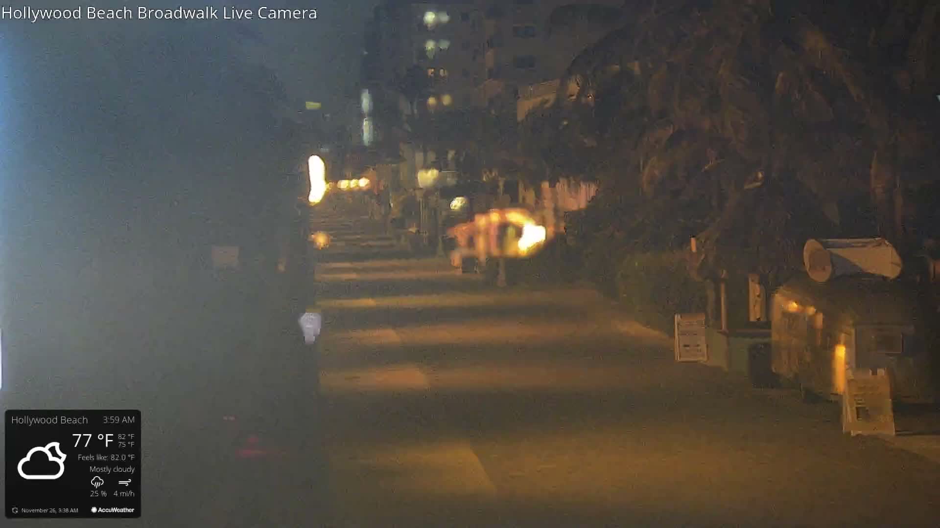 A dimly lit outdoor path at night, flanked by dark trees and a small illuminated concession-like stand on the right, with faint building lights visible in the distance, under mostly cloudy skies at 77°F.