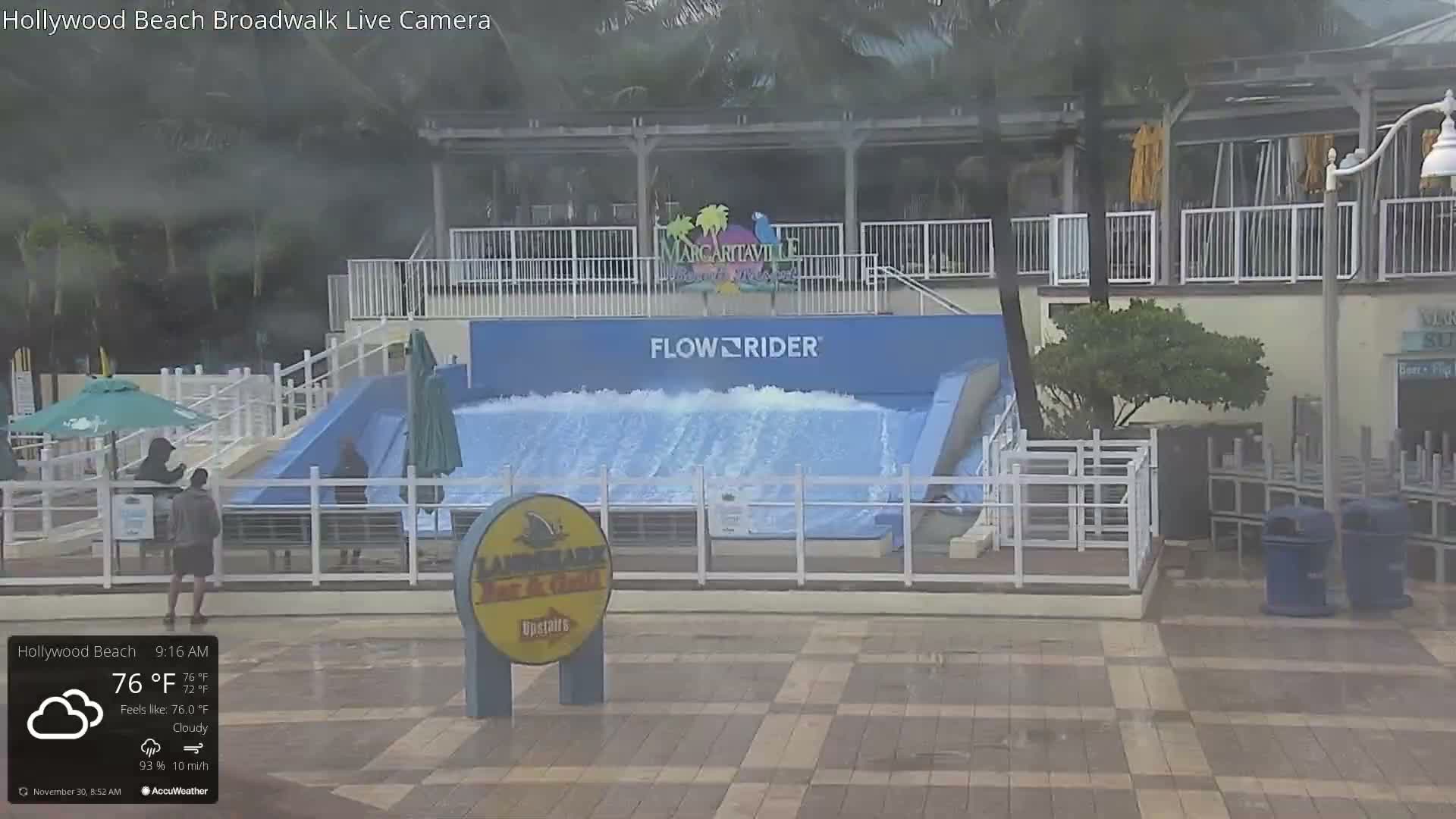 An outdoor wave simulator with a continuous sheet of water is the focal point of a bustling plaza where people watch and walk, set amidst tropical trees and resort architecture, under mostly cloudy skies with a temperature of 72°F.